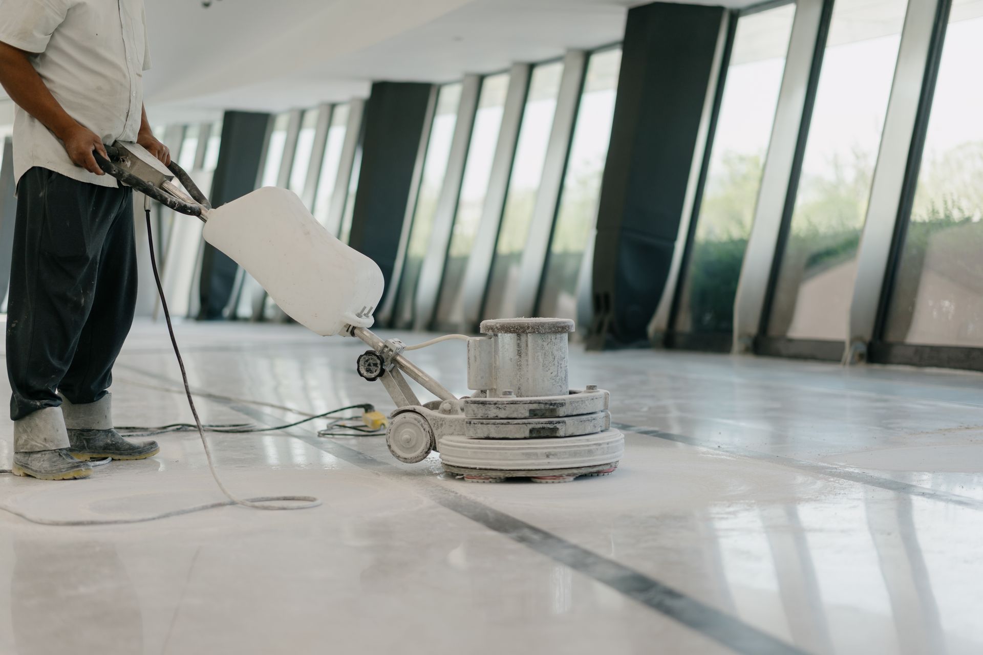Person cleaning a hallway floor with a floor buffer, large windows in background.