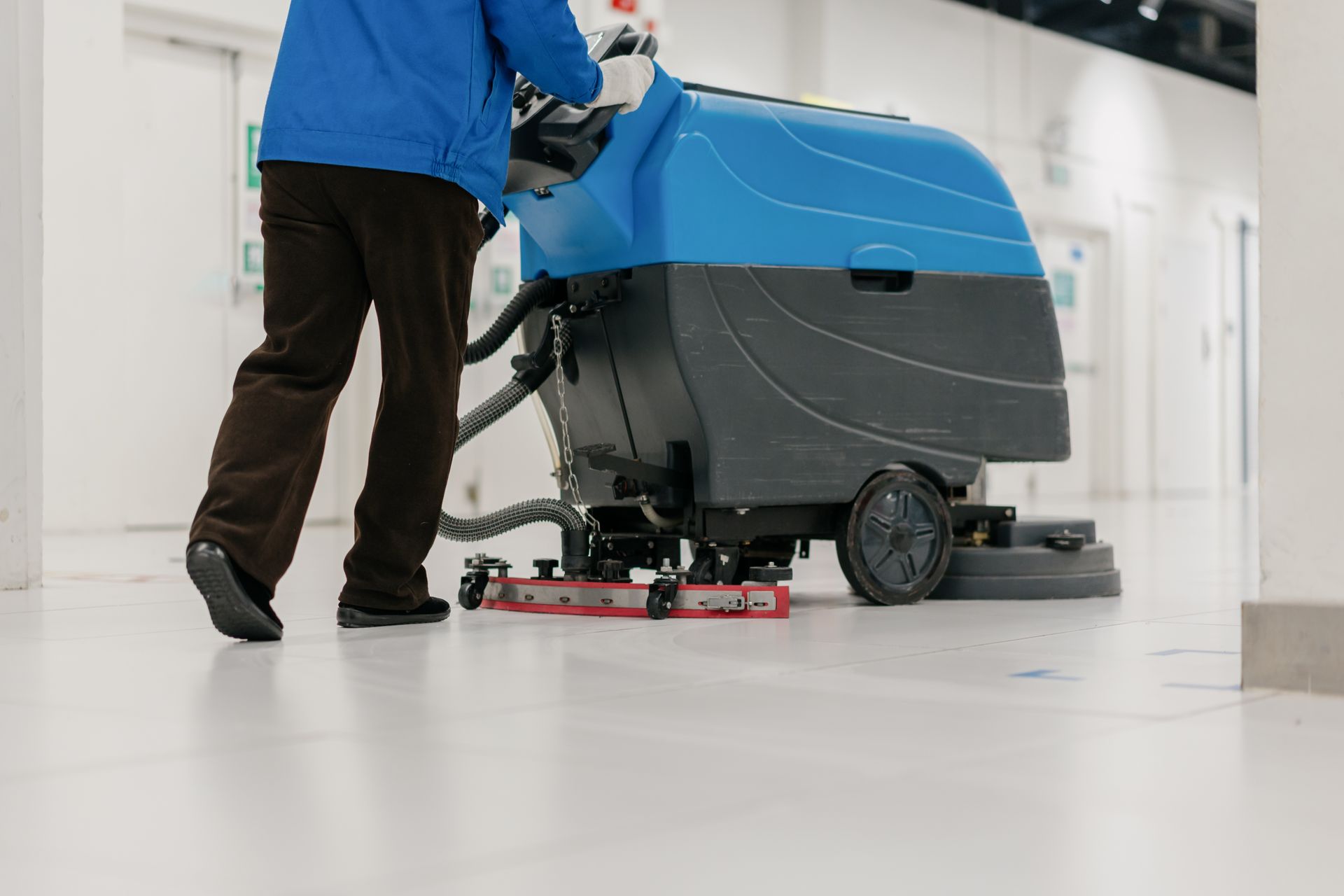 Person operating a blue and black floor cleaning machine in a white-floored hallway.