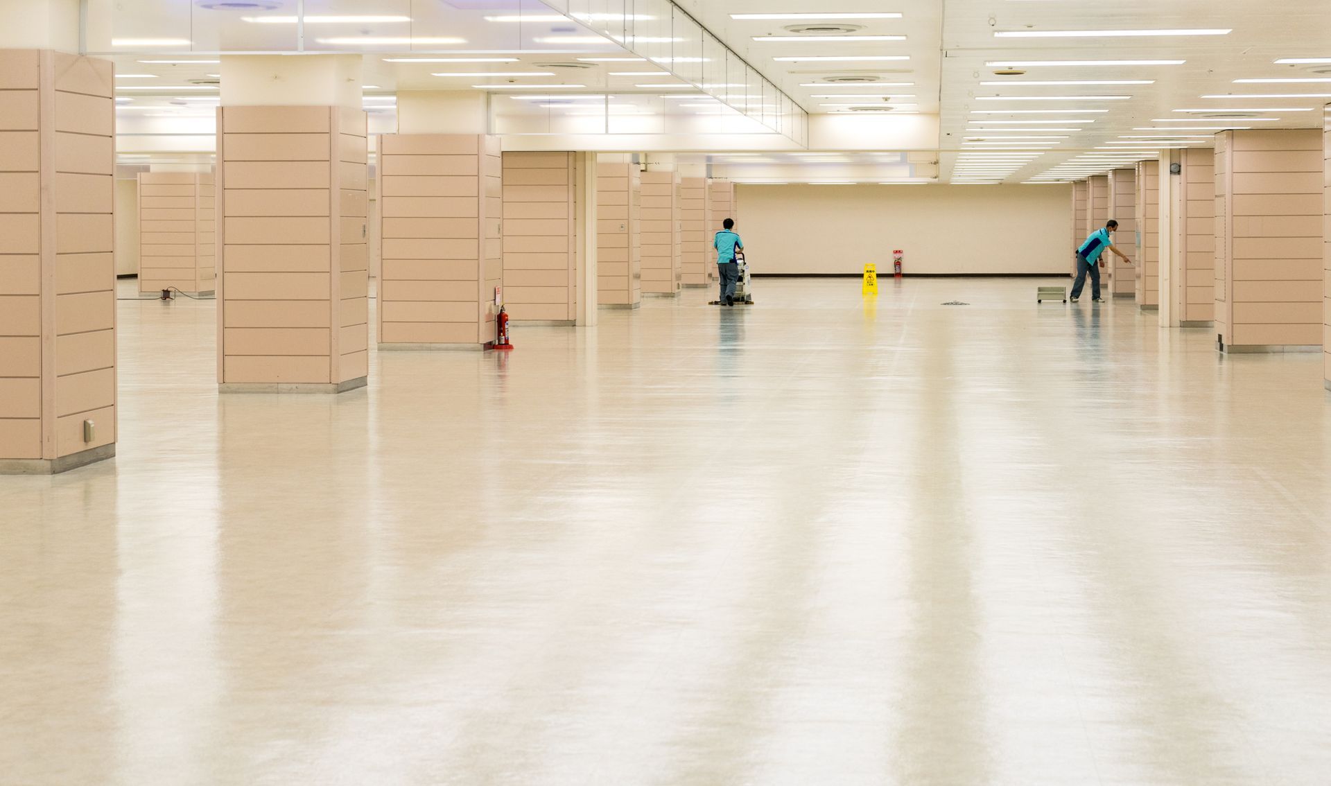Two people mopping a large, empty, light-colored, tiled floor in a building. Beige pillars.