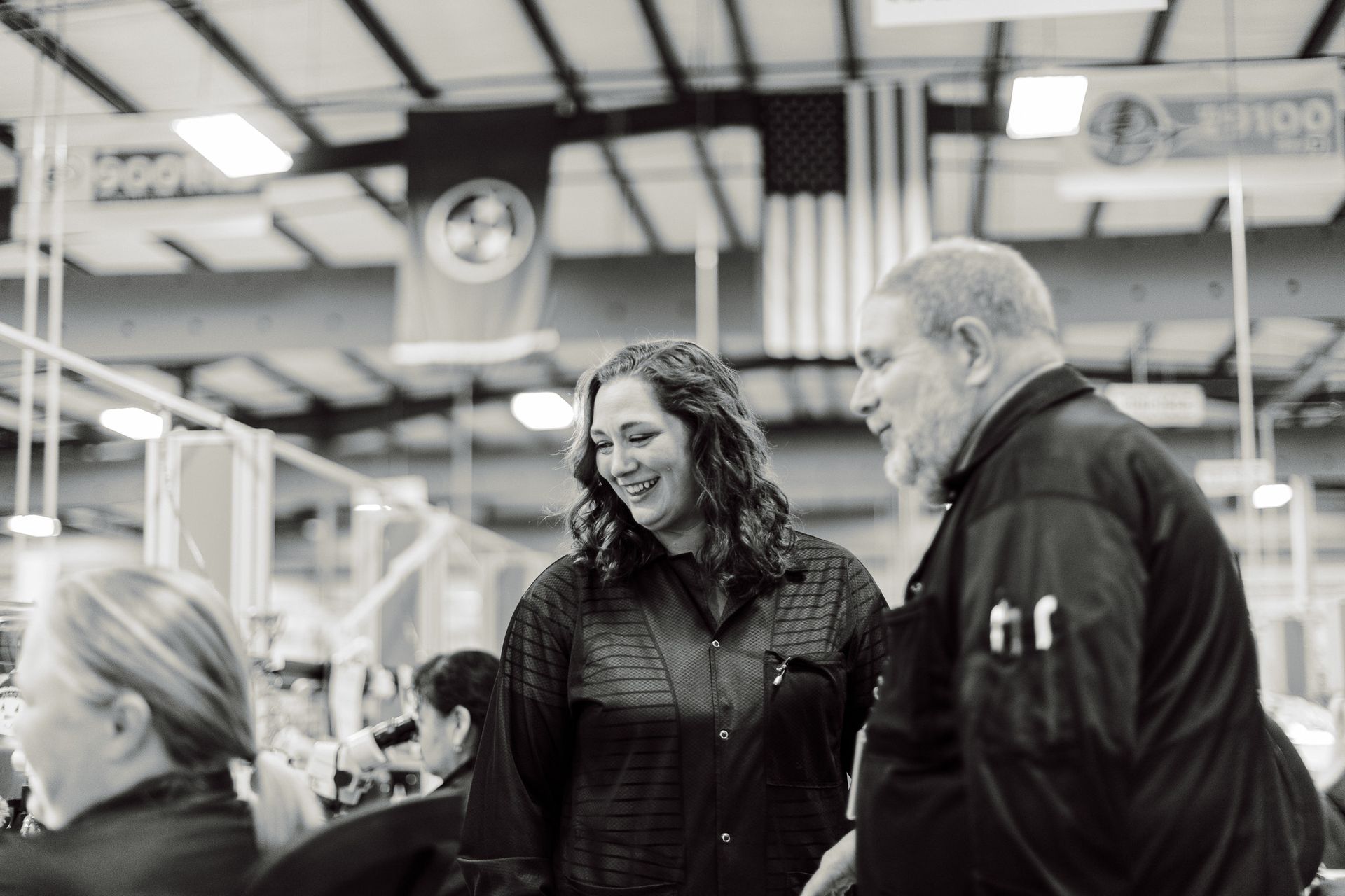 Woman and man in an industrial setting, smiling and talking. American flag and factory equipment visible.