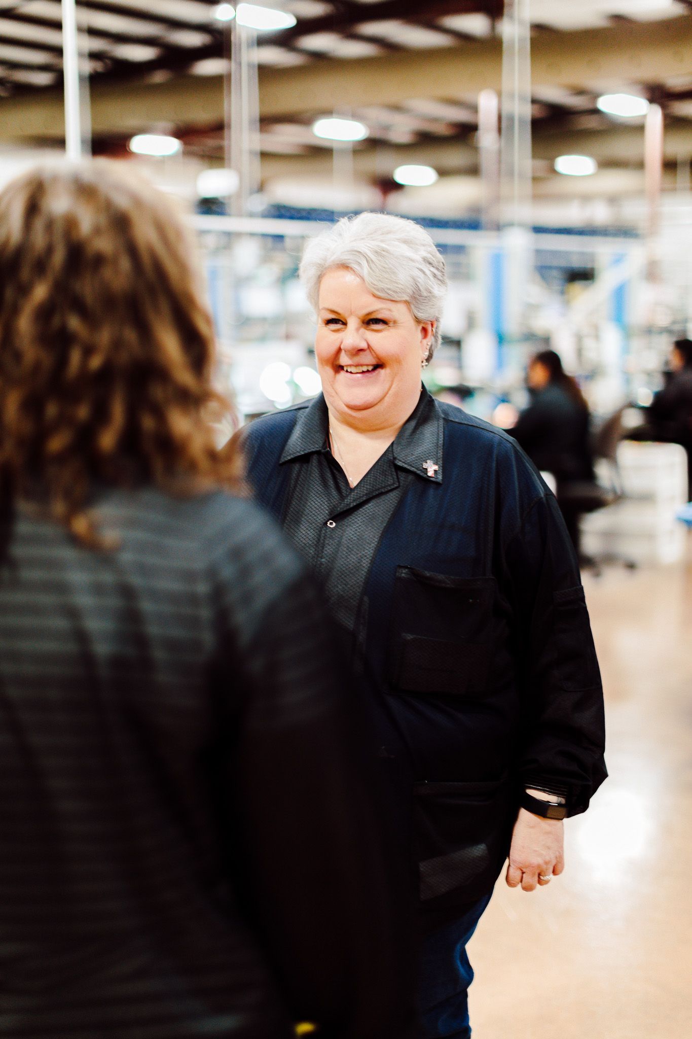 Woman with short grey hair smiles in a factory. She wears a navy blue shirt and is talking to someone.