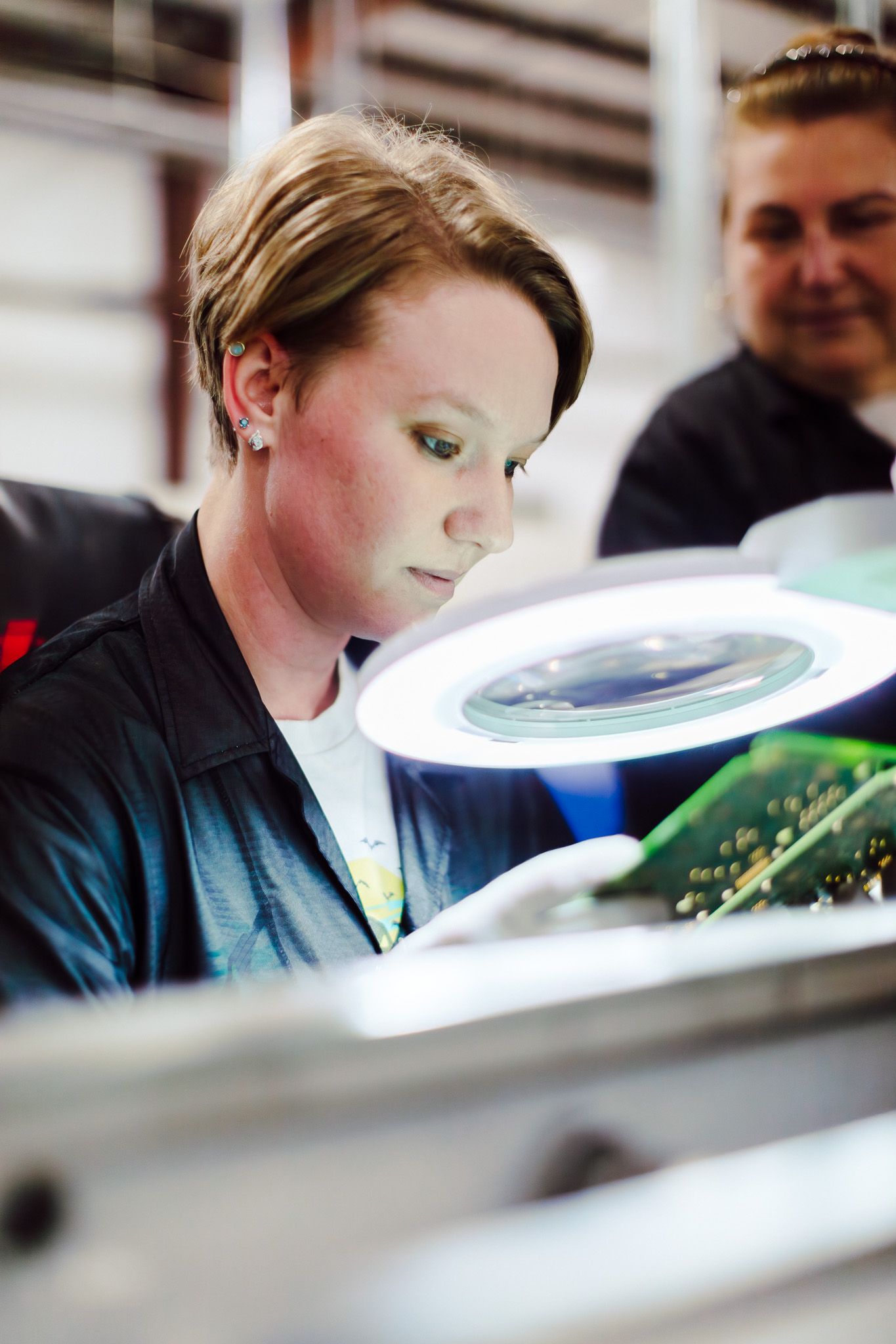 Woman inspecting a circuit board under a magnifying lamp in a manufacturing setting; another person in the background.