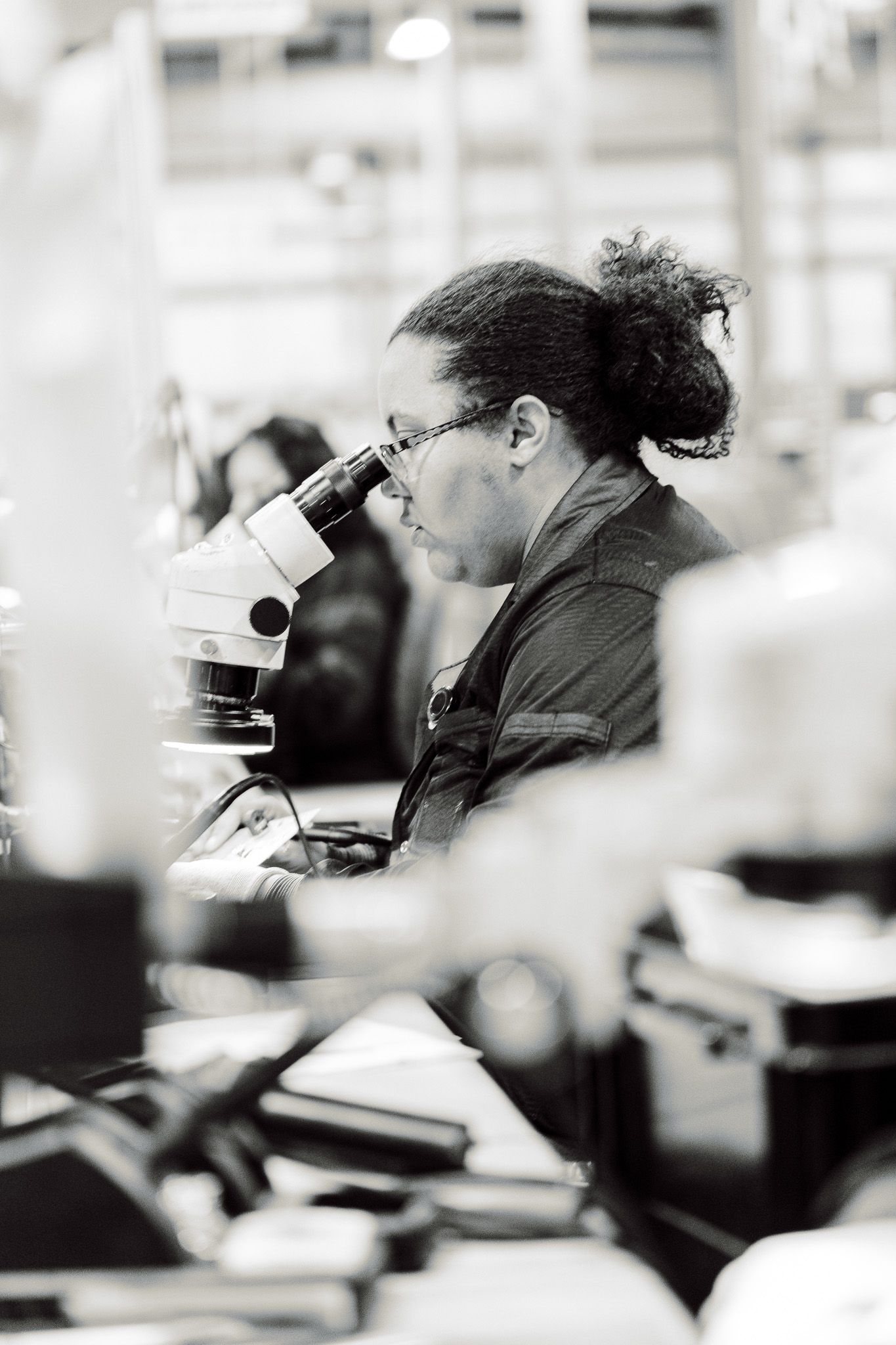 Woman with dark hair, wearing glasses, looking into a microscope in a lab.