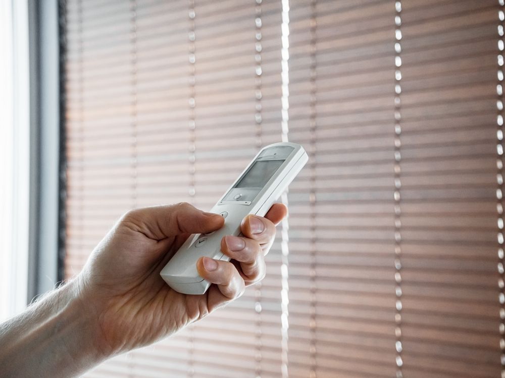 Hand Holding a White Remote Control in Front of Window Blinds — Wares Blinds, Screens & Awnings In Mount Larcom, QLD