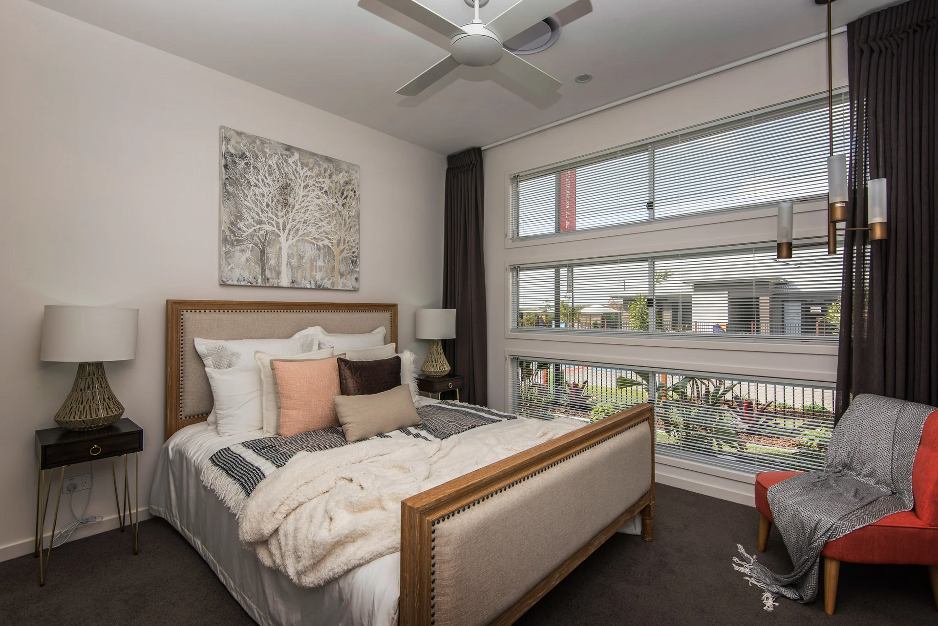 Bedroom with bed, art, window, red chair, lamp. Neutral colors, natural light.