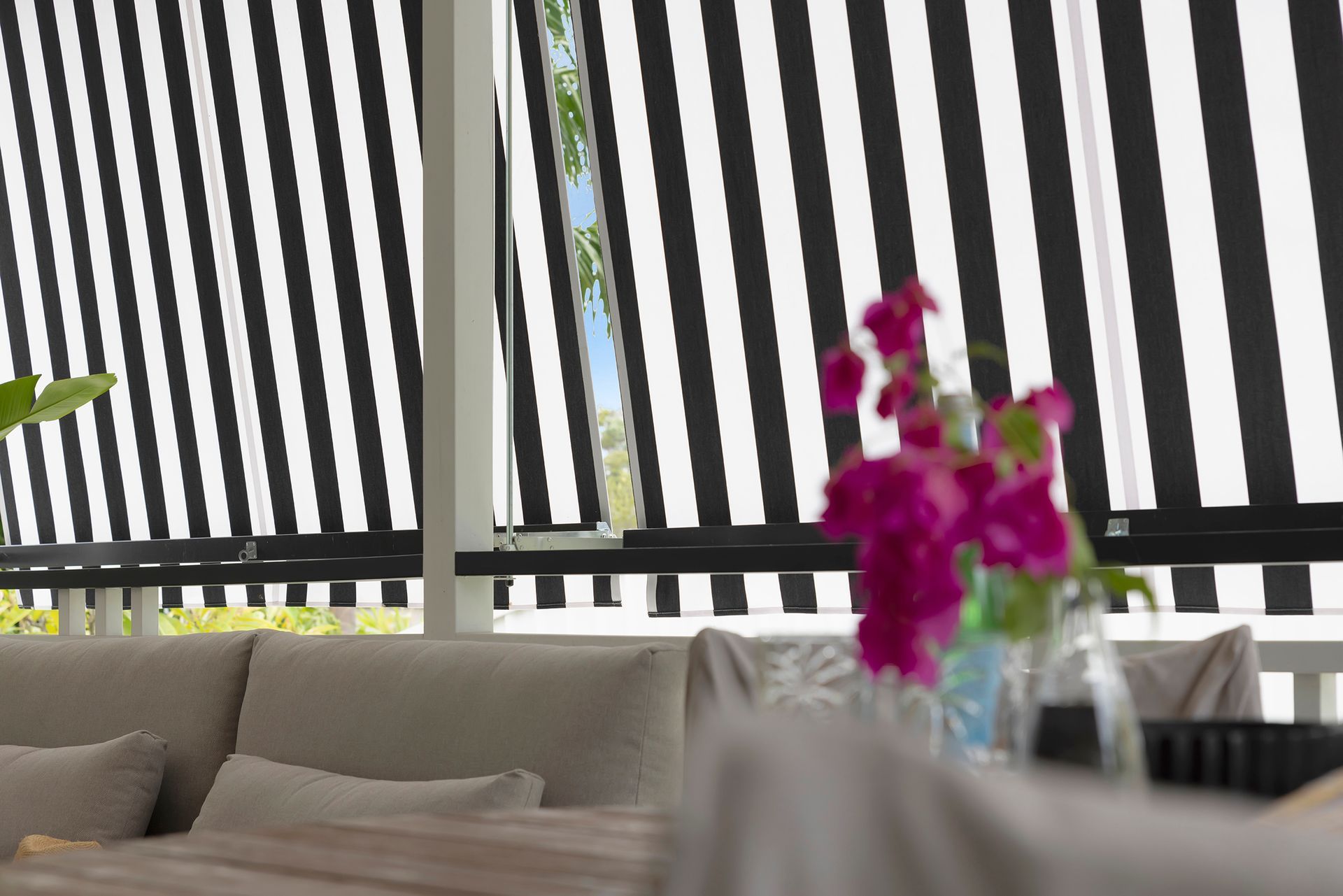Black and white striped outdoor blinds partially cover a room with a beige sofa and pink flowers.