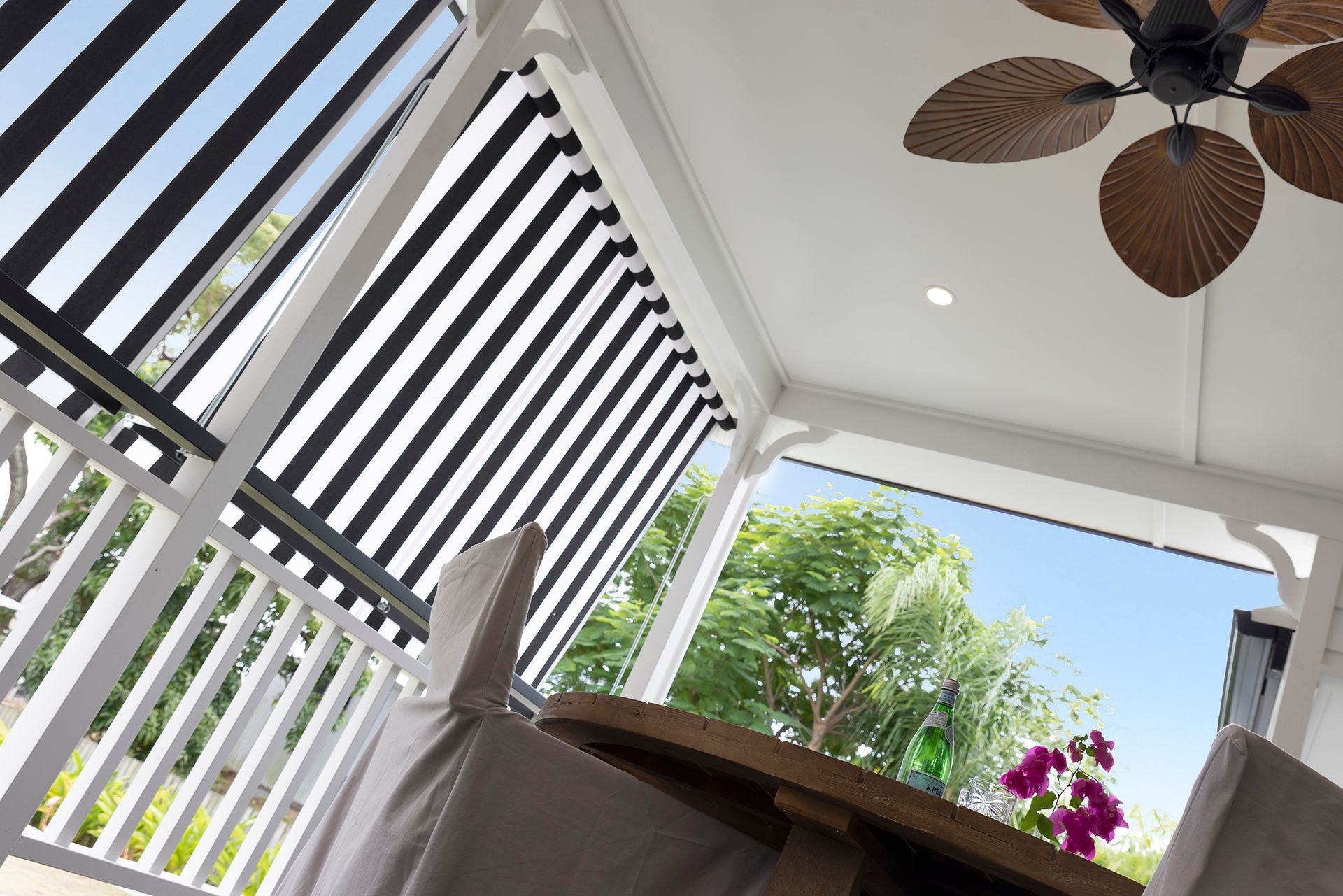 Outdoor veranda with black and white striped shade, ceiling fan, and table with chairs; view of trees.