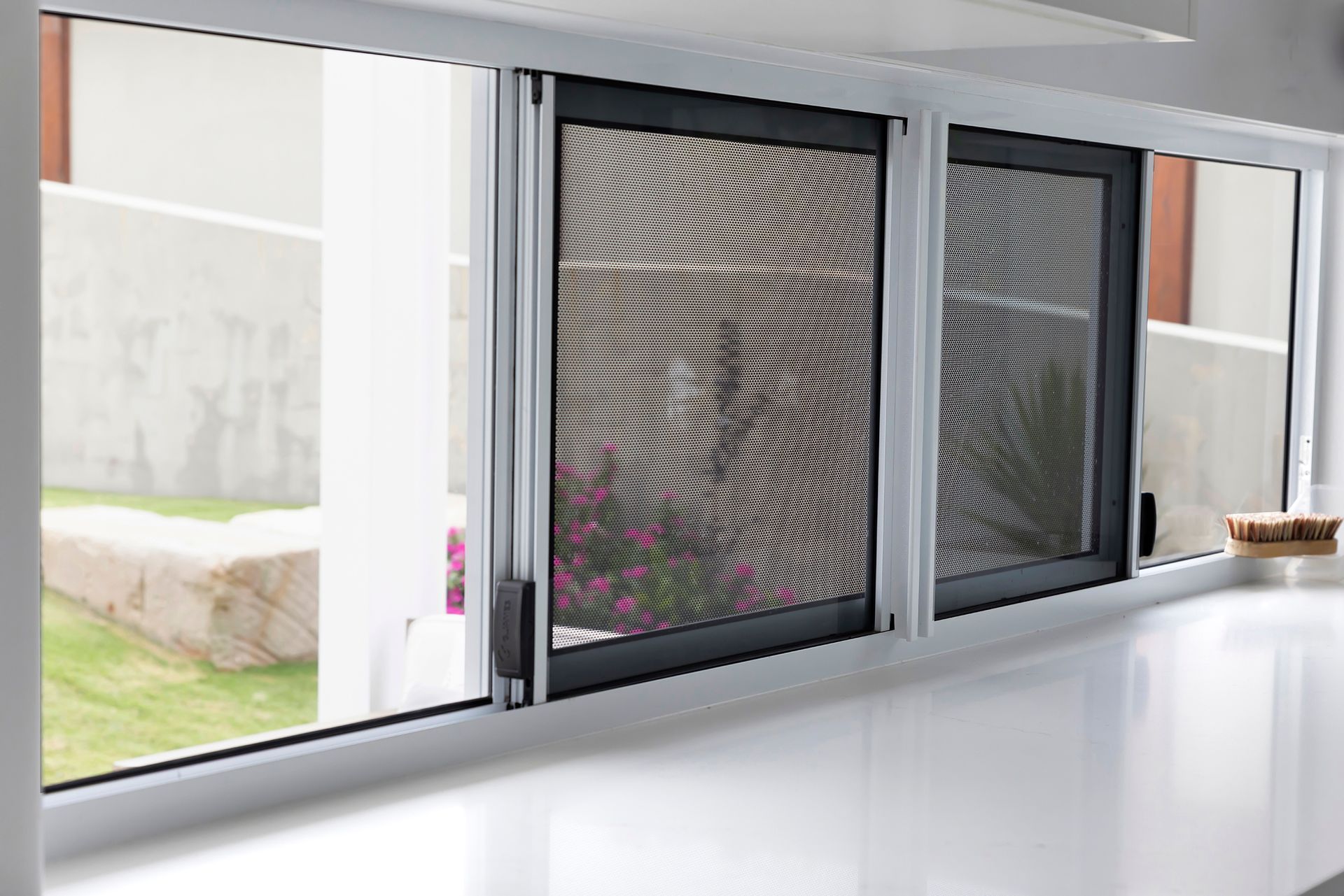 Sliding windows with black screens and white frames, on a white countertop, overlooking a yard.