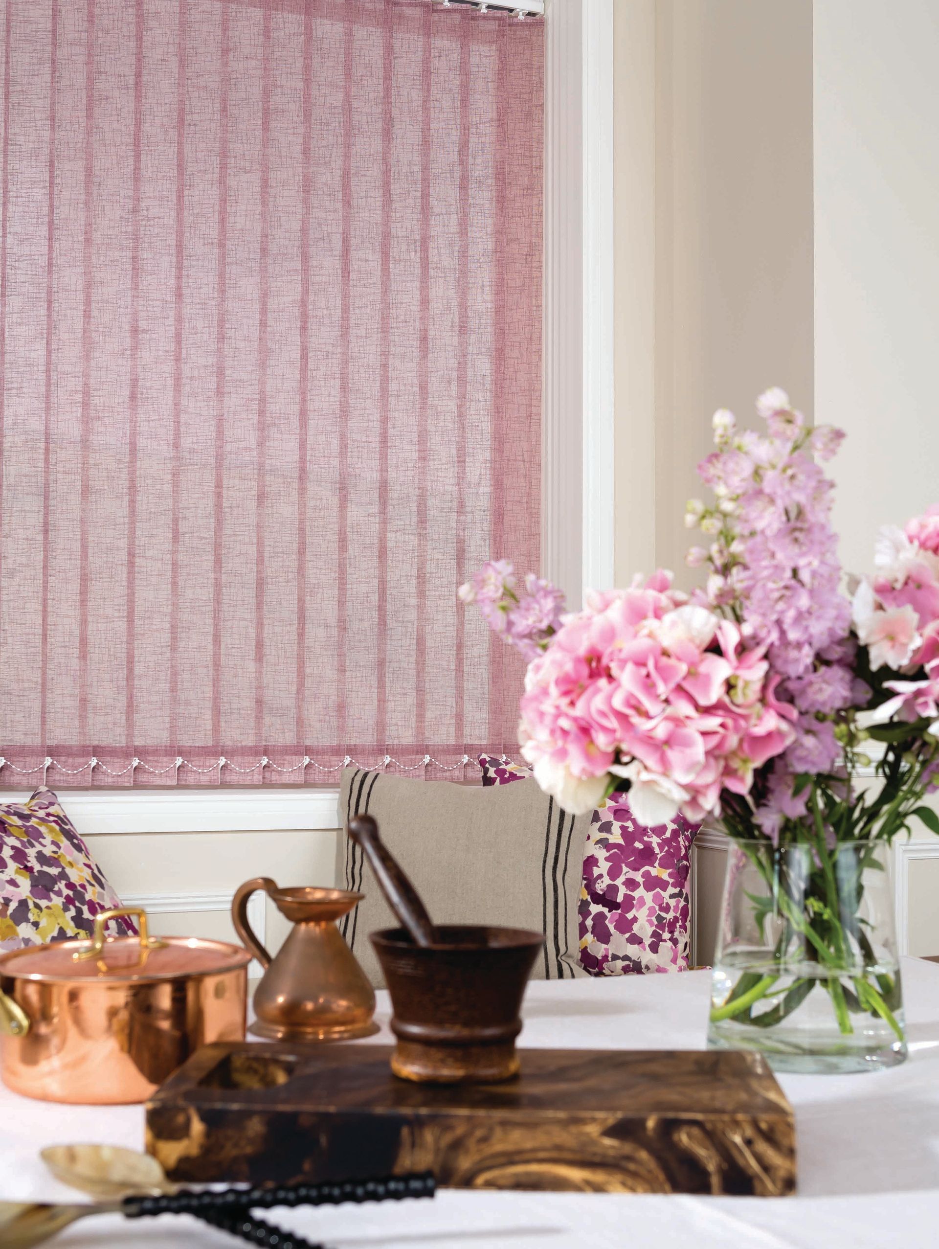 Pink vertical blinds next to a table setting with flowers, copper pots, and a wooden cutting board.