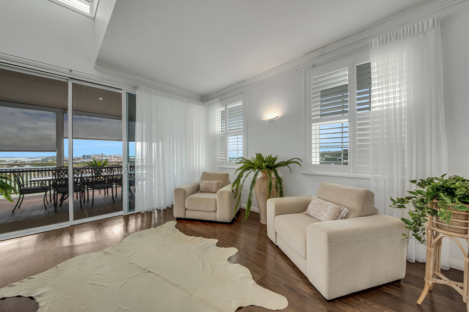 Cozy living room with cream armchairs, sheer white curtains, wooden floors, and a balcony view.