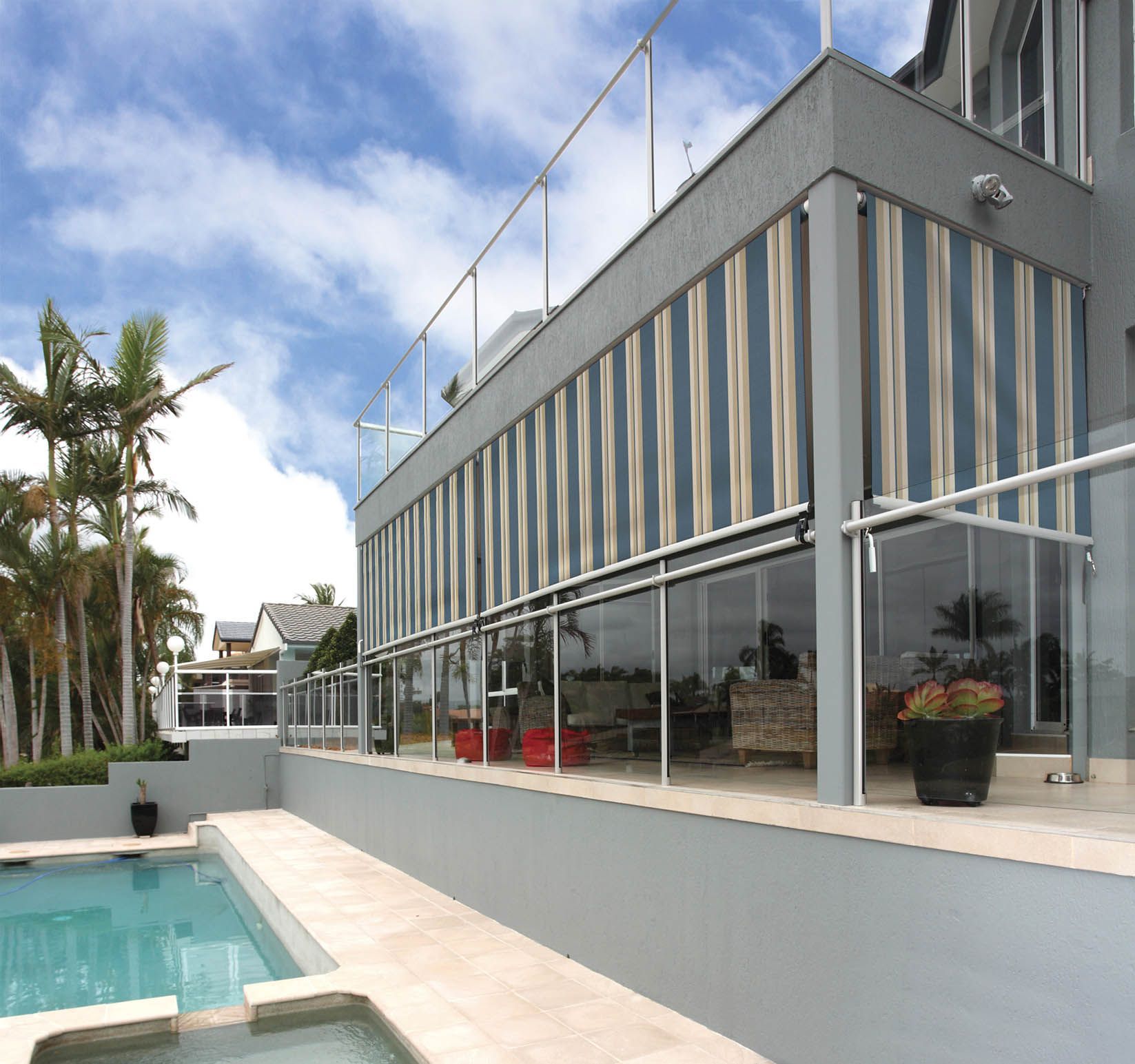 Modern home exterior with pool. Striped awnings, glass railings, and a blue sky.