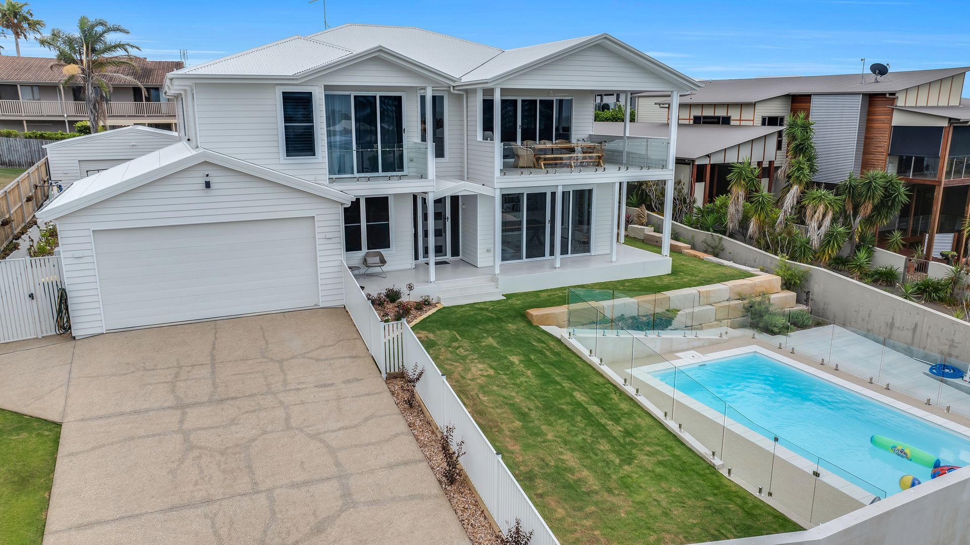 White two-story house with balcony, garage, and pool in front yard; green lawn and blue sky.