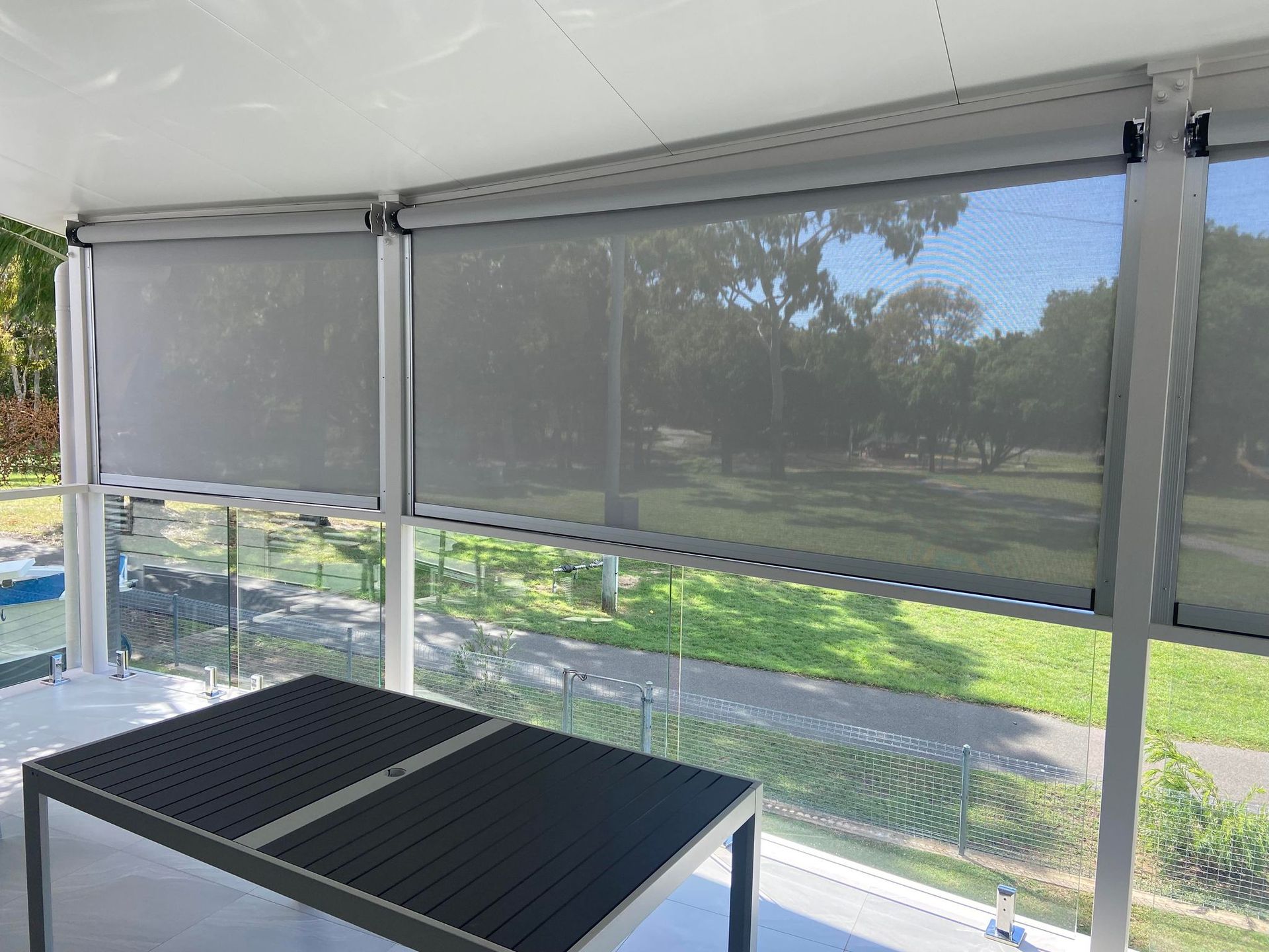 Exterior view of gray roller blinds partially covering windows overlooking a park with a table in the foreground.