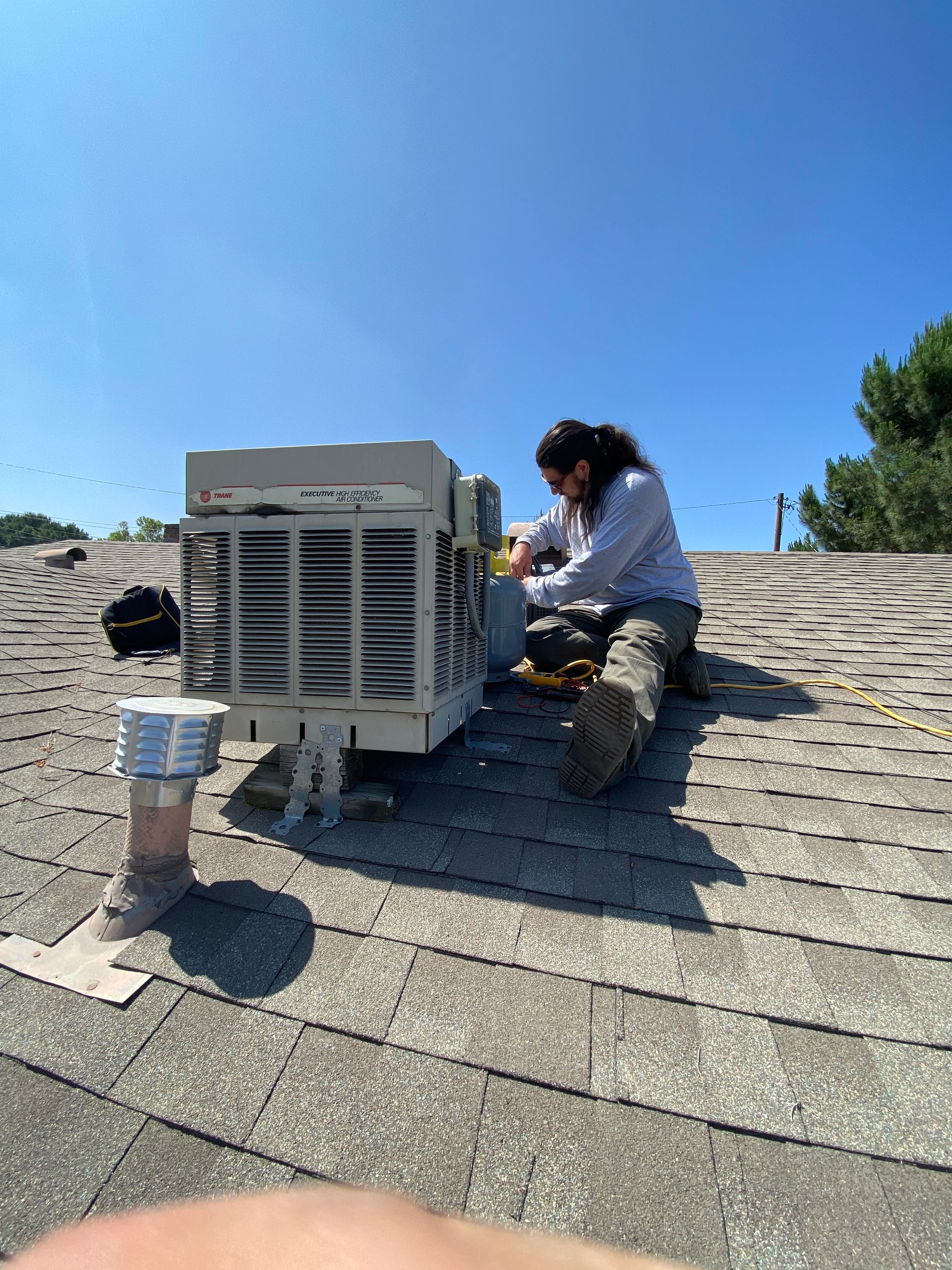 A man is sitting on top of a roof working on an air conditioner.