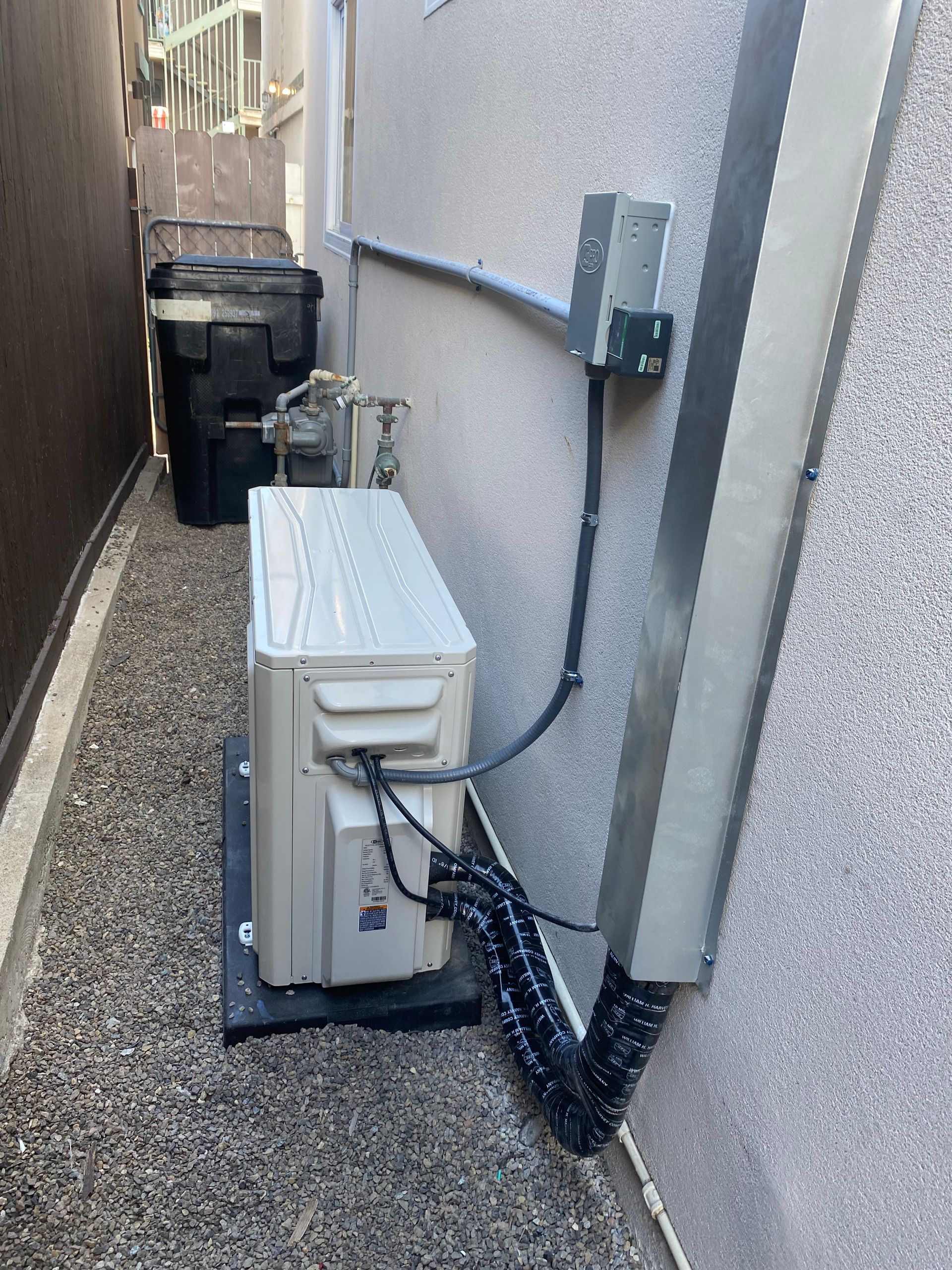 An air conditioner is sitting on the side of a building next to a trash can.