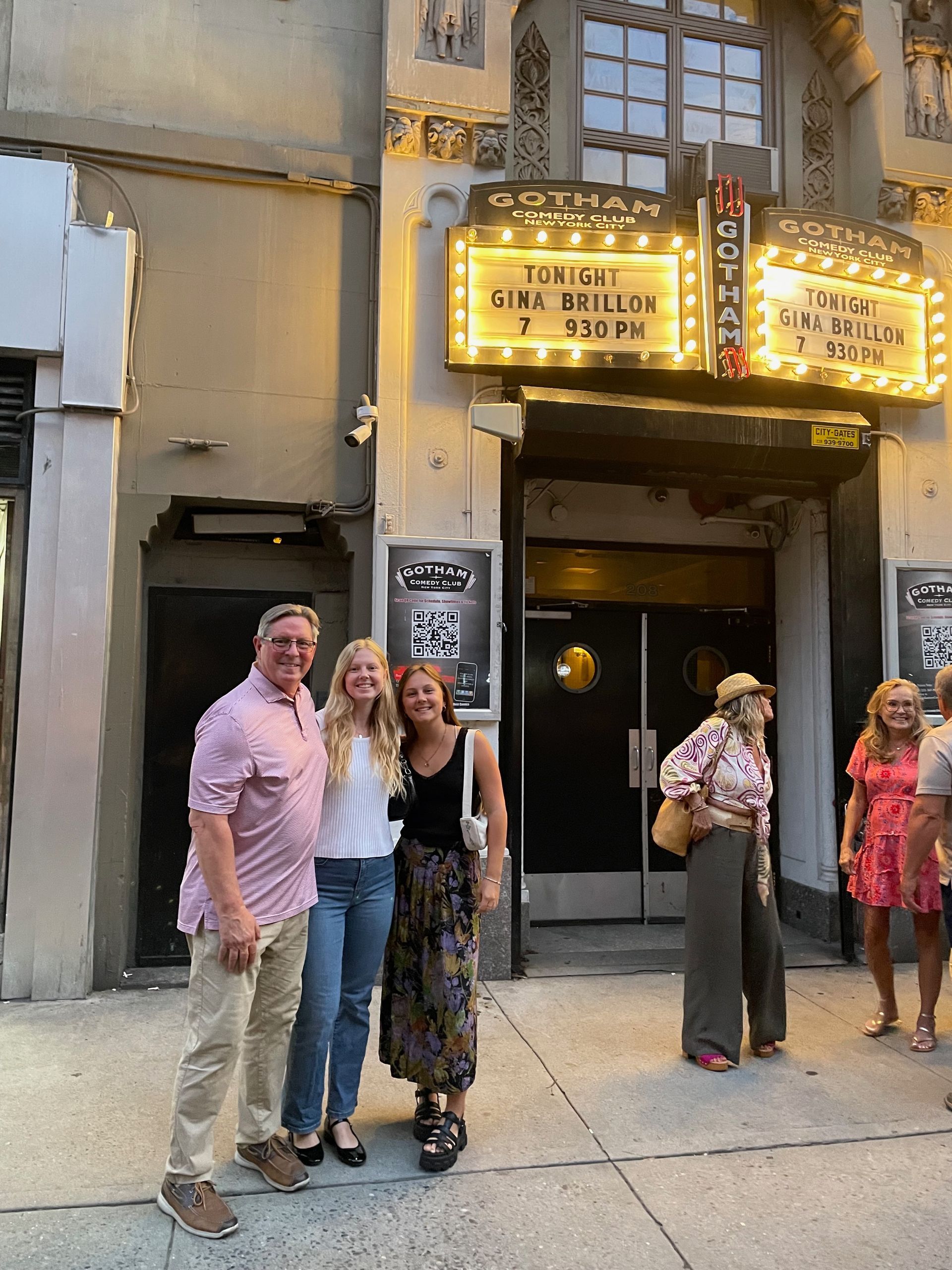 People pose in front of a theater. A marquee reads "Gotham." The person on the left wears pink and khakis. Others wear jeans, black tops, and floral prints.