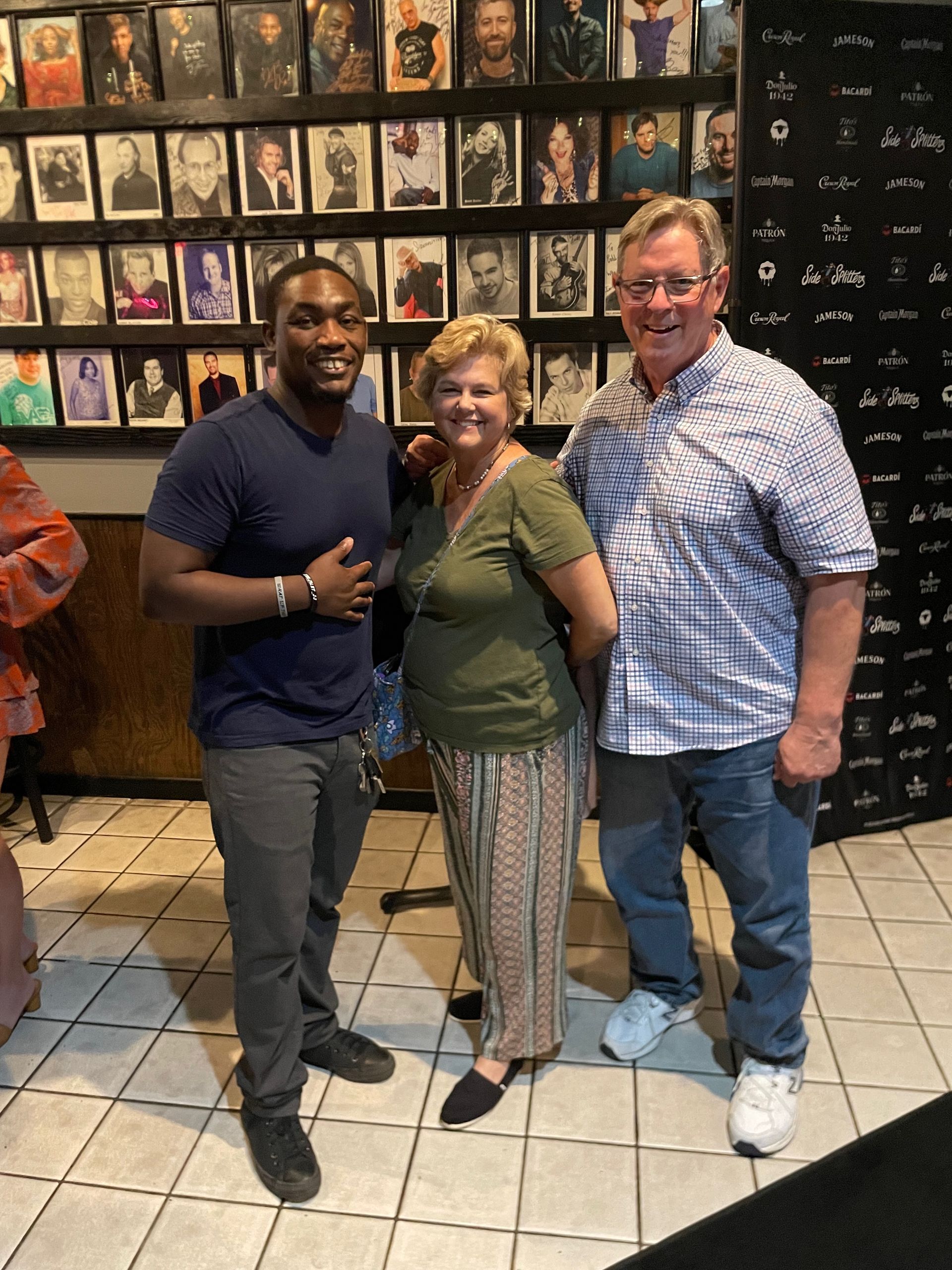 Three people pose for a photo in front of a wall of portraits. The person on the left wears a blue shirt and grey pants; the middle person wears a green shirt and patterned pants; the person on the right is wearing a blue and white checkered shirt and jeans.