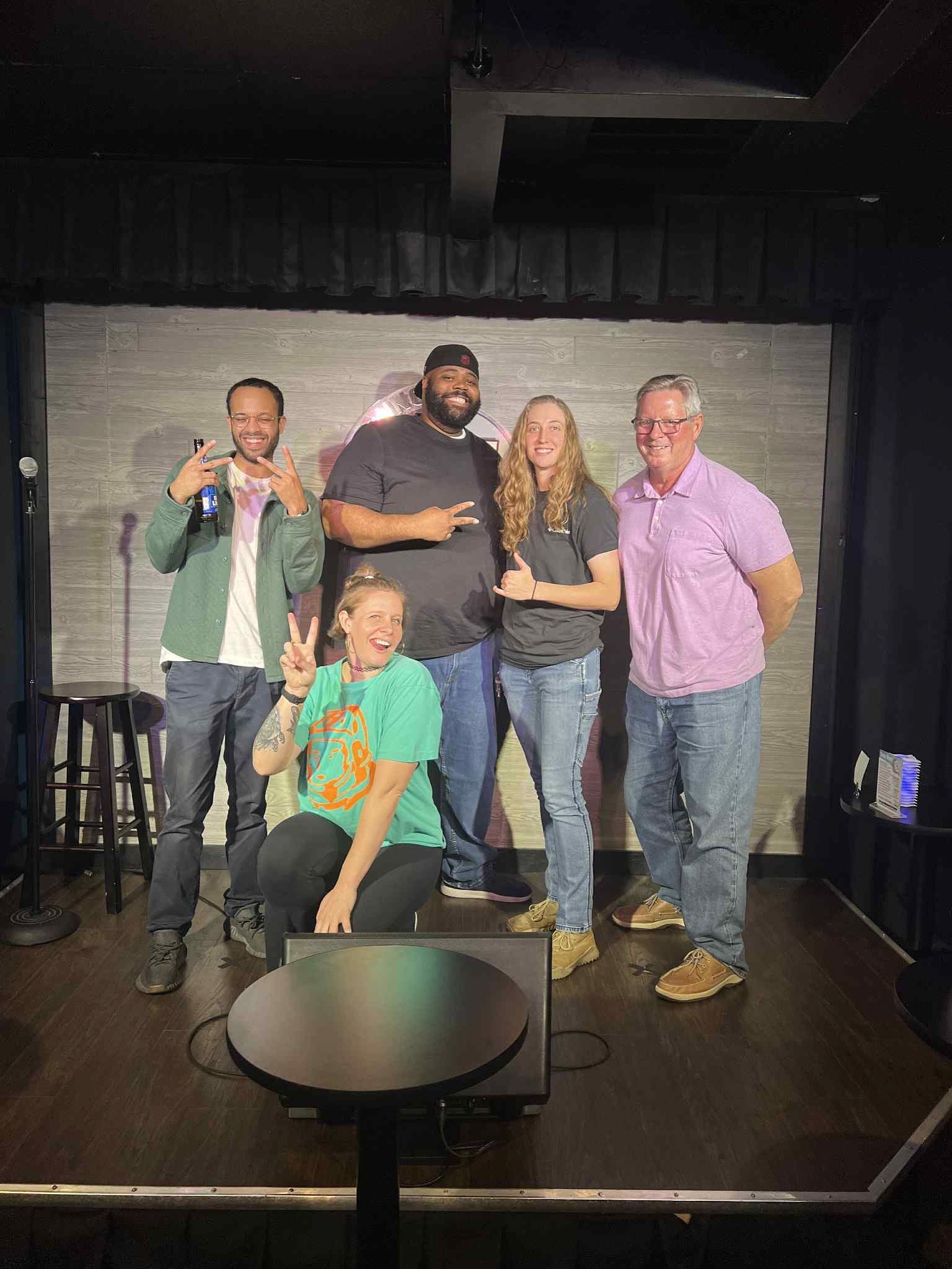 Five people on a stage. Two standing and three posing. They are smiling and making peace signs.