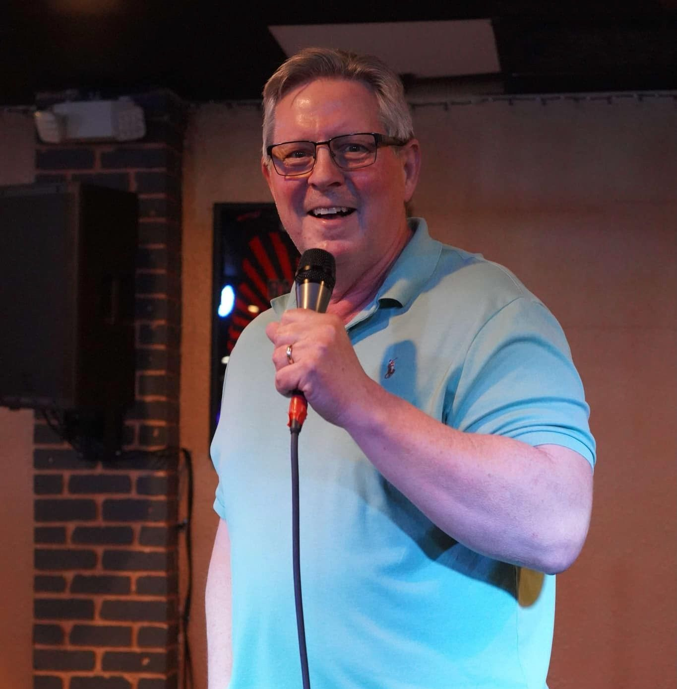Man in light blue shirt holding microphone, smiling, on stage. Brick wall backdrop.