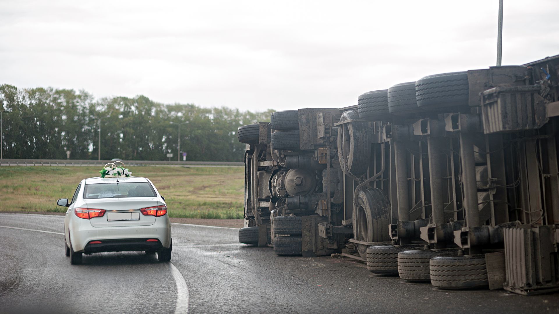 A car drives near a semi-truck that has rolled over on a wet highway.