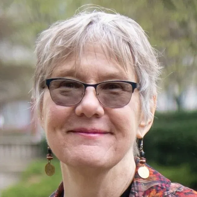 Woman with short gray hair and glasses, wearing earrings, smiling outdoors.