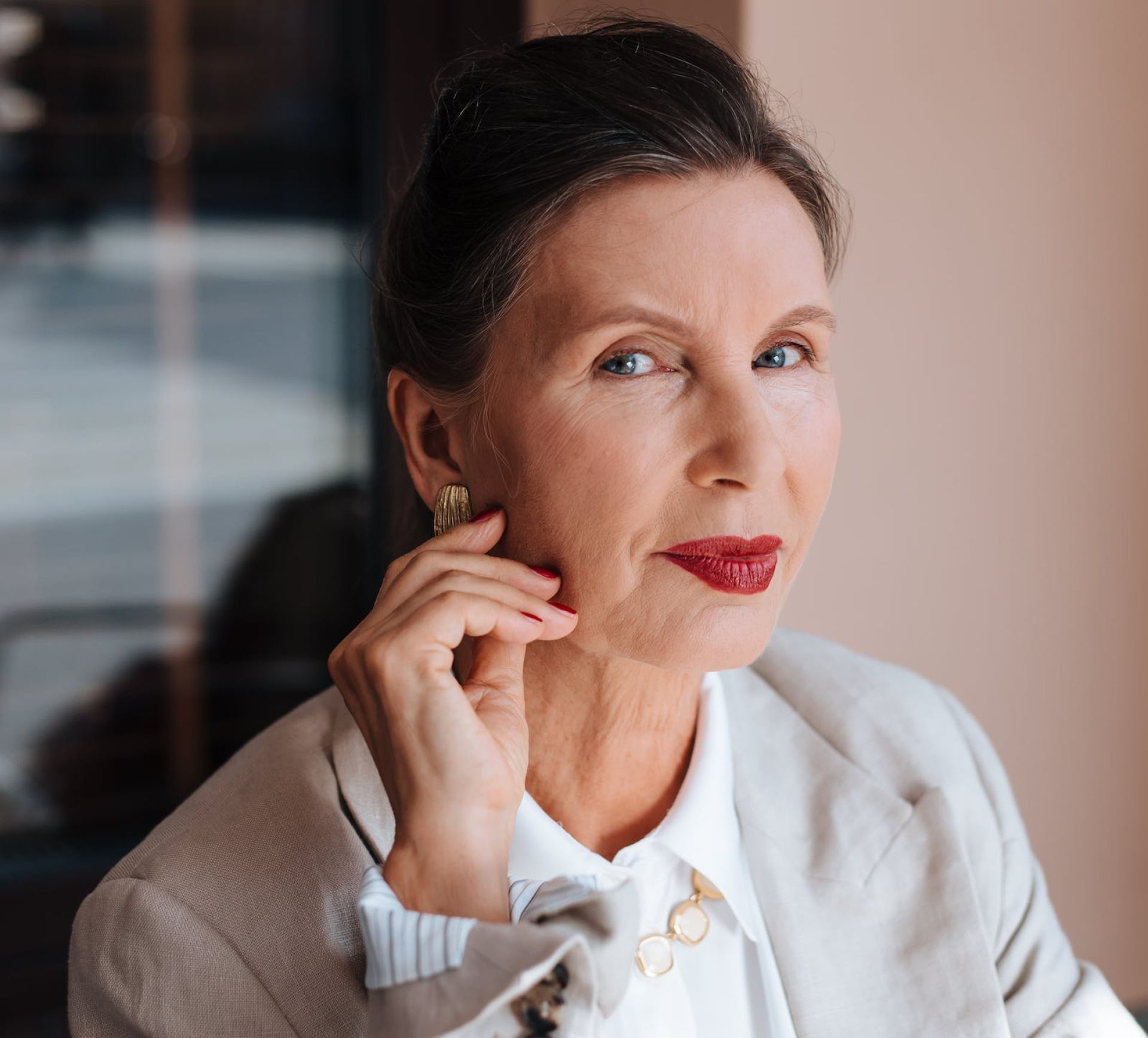 Woman with red lipstick, resting hand on her chin, wearing a blazer and gold earrings.