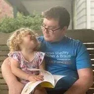 Man in blue shirt reading to a child with curly hair outdoors.