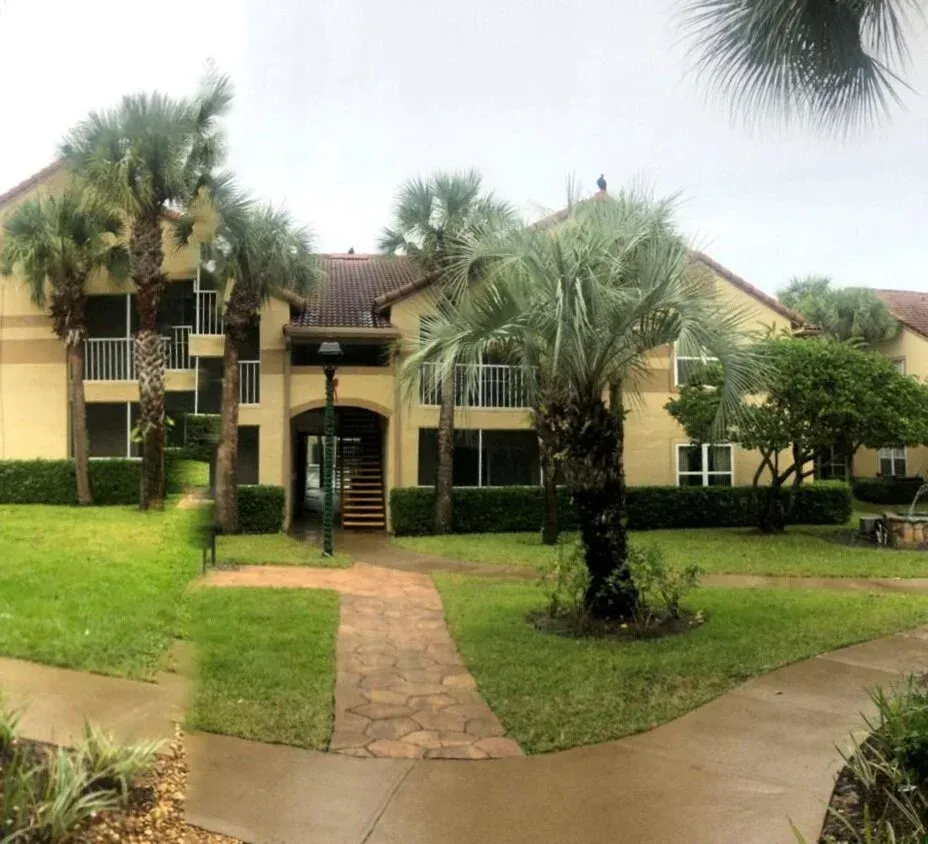 Apartment building with beige walls, palm trees, and a brick path leading to the entrance.