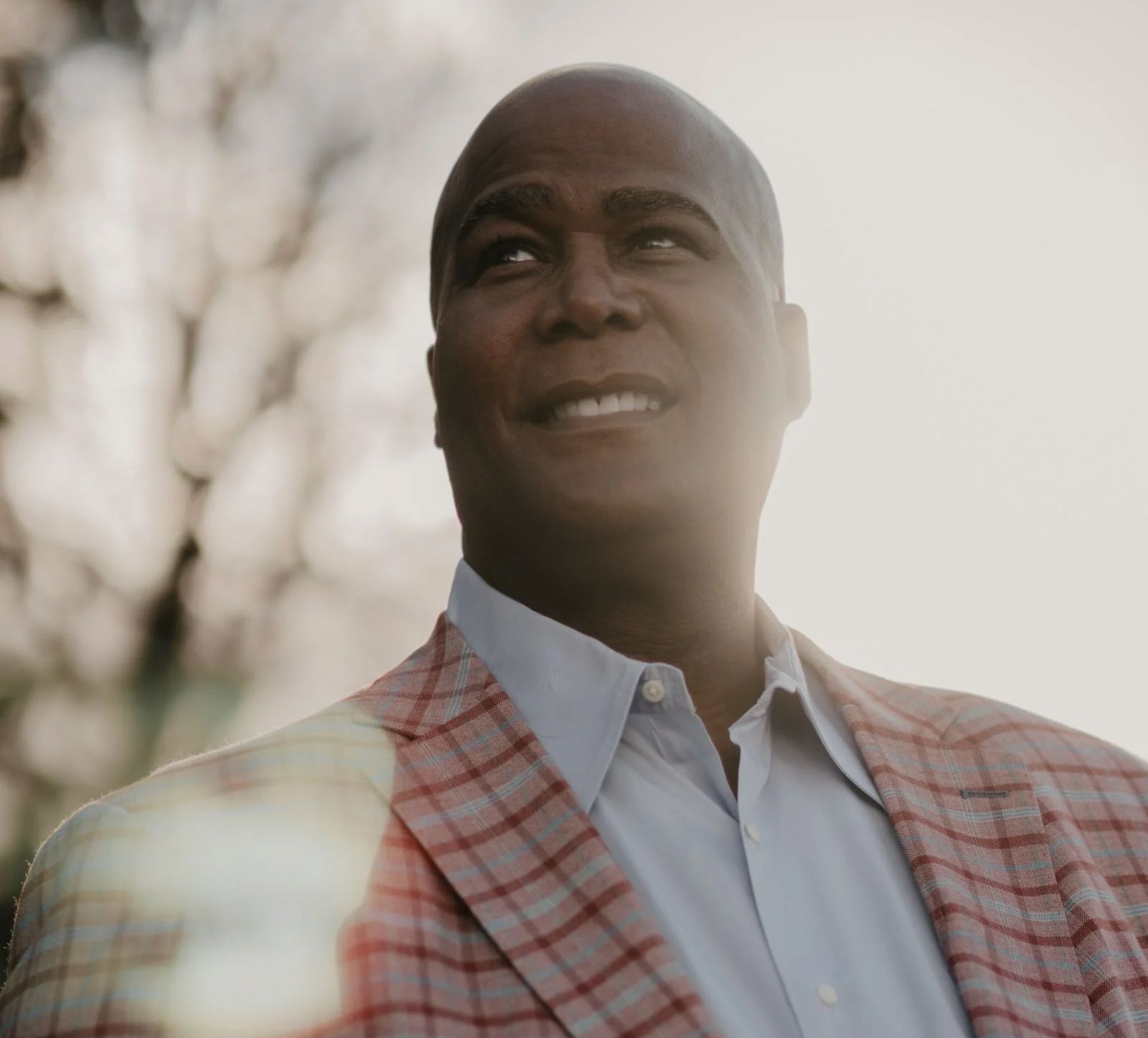 Smiling Black man in a plaid blazer and light blue shirt, looking upwards against a blurred sunlight background.