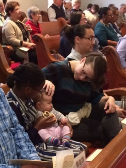 People in a church. A woman holds a baby, another woman leans over them smiling.