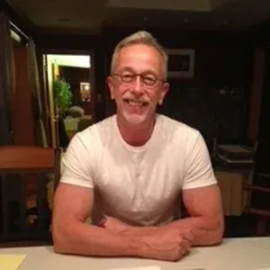 Man in white t-shirt smiles, arms crossed at a table, glasses. Indoors, neutral background.