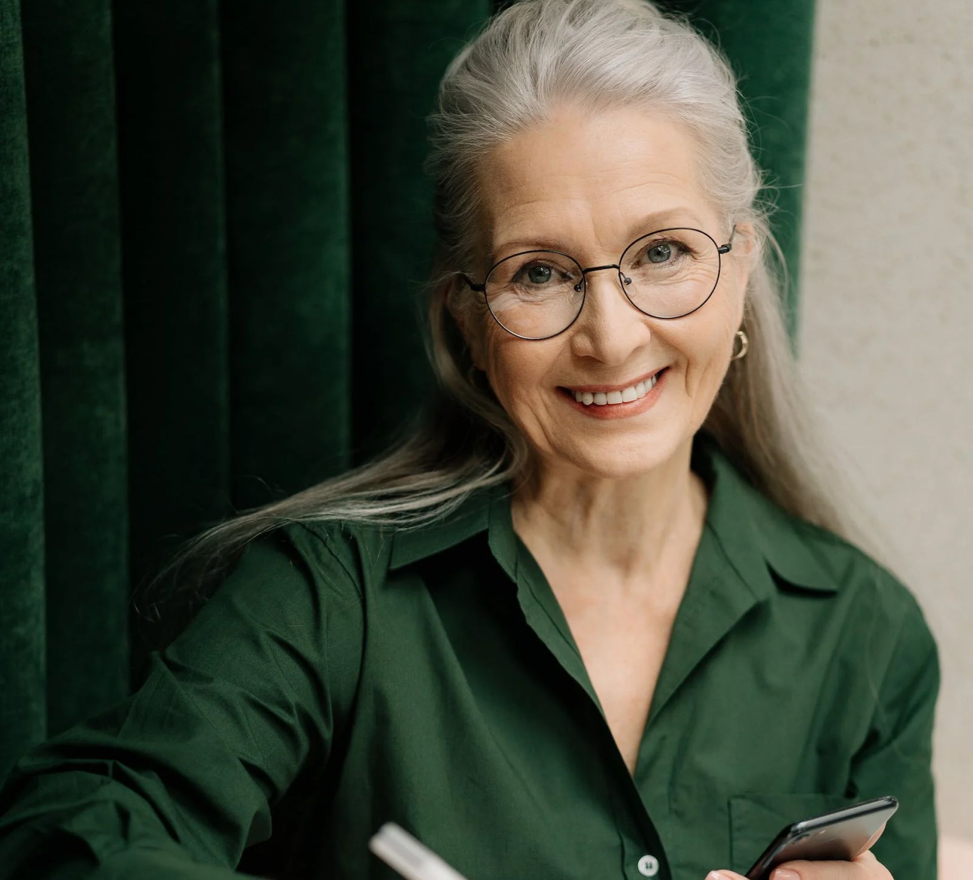 Smiling older woman with glasses in a green shirt, holding a phone and a pen.