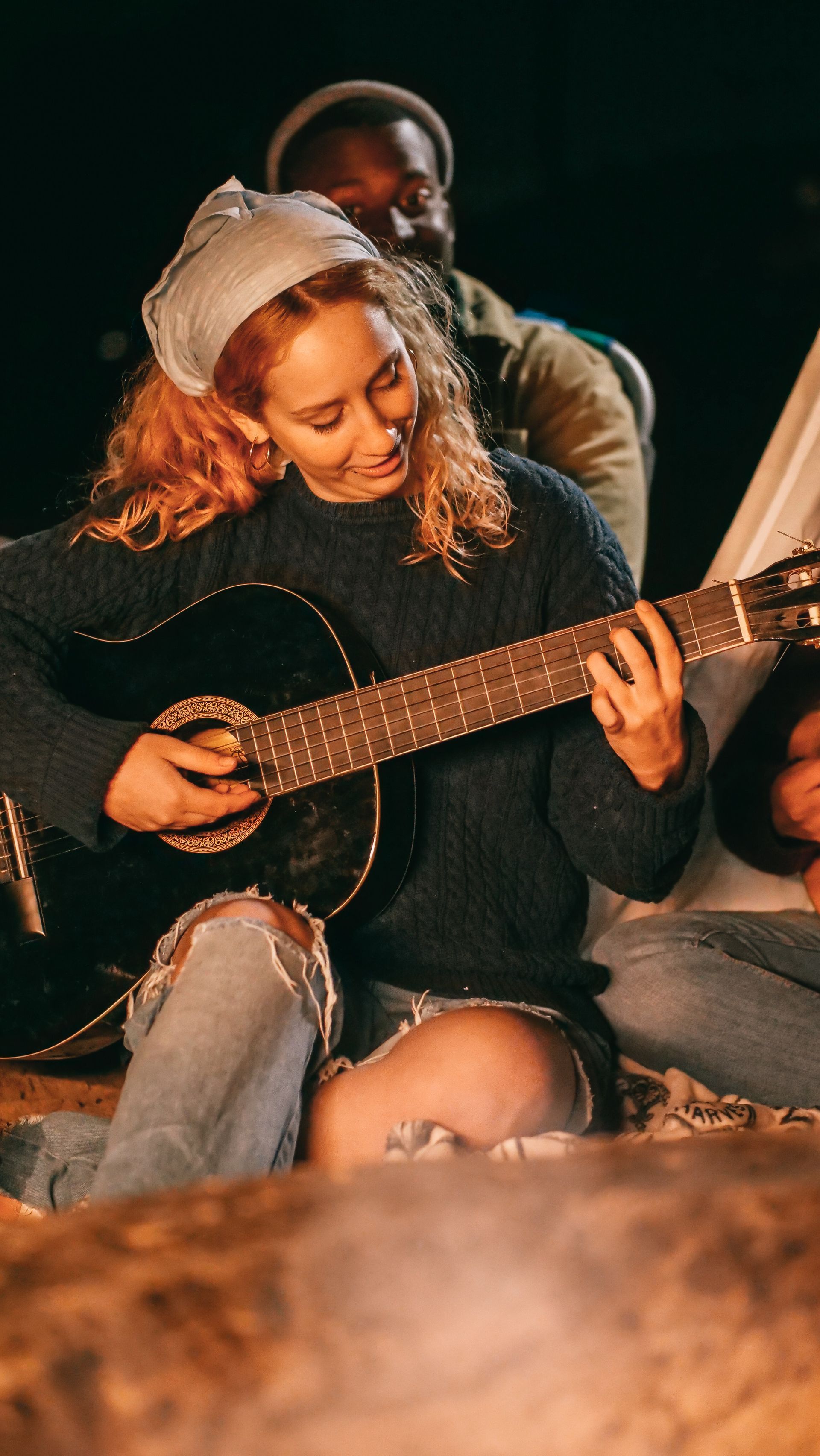 Woman with red hair plays guitar by campfire with friends at night.