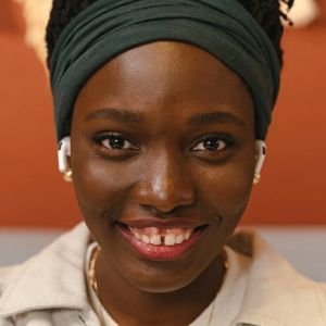 Close-up of Black woman with dark hair, a green headscarf, and AirPods smiling, wearing a beige shirt.