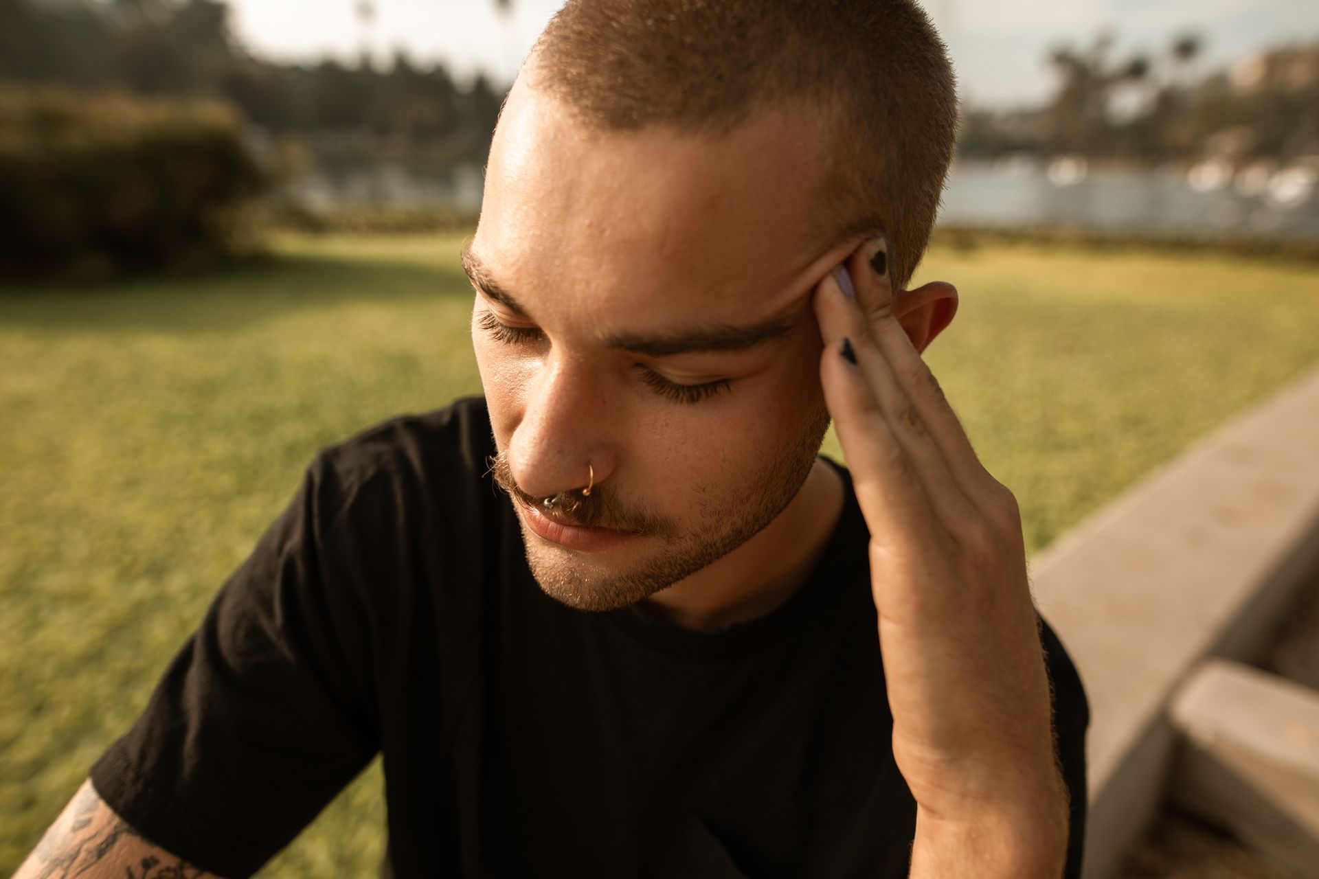 Man with a nose ring and black nail polish touches his forehead, looking down in a park.