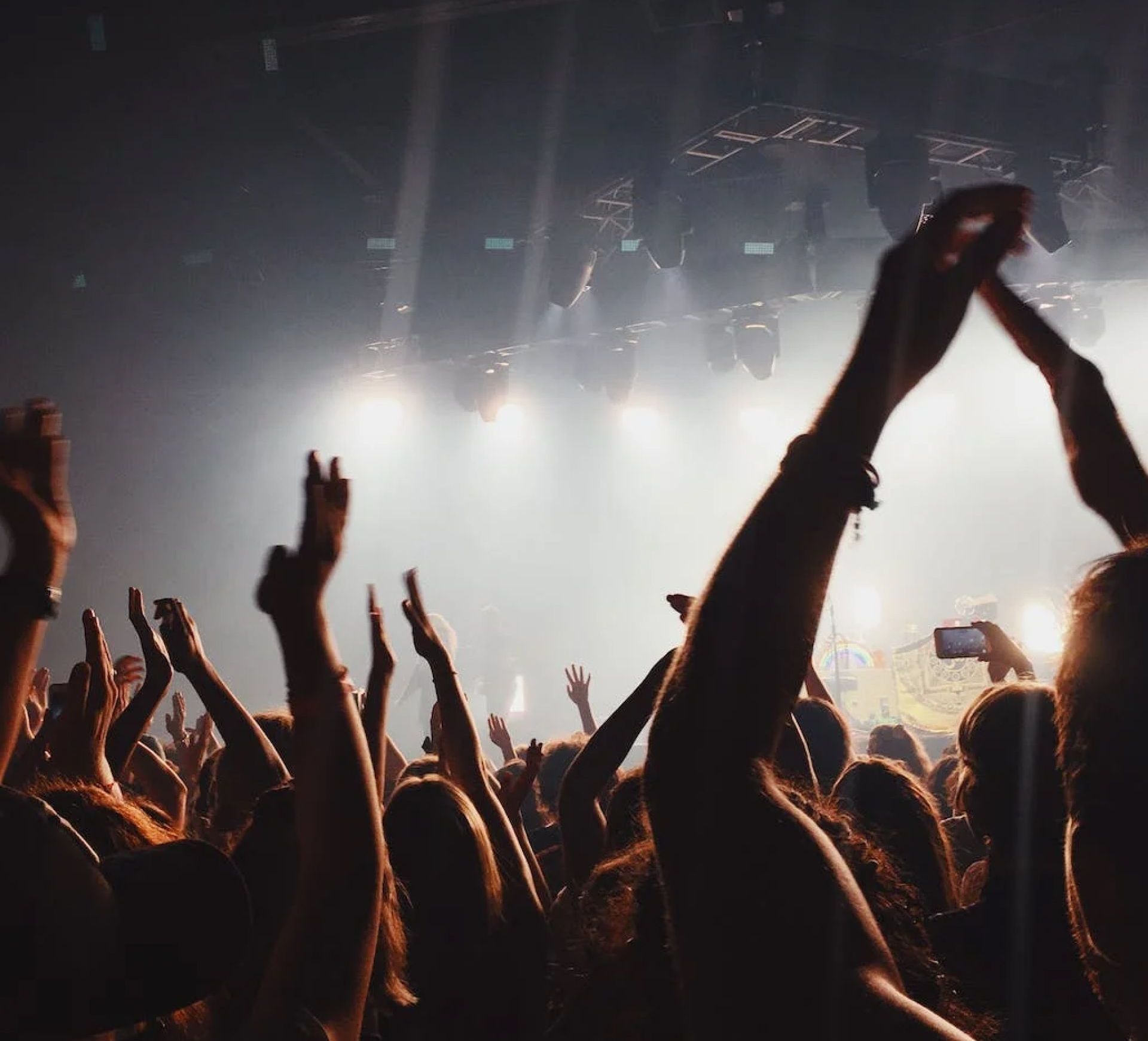 Concert crowd with arms raised in the air, illuminated by stage lights and smoke.