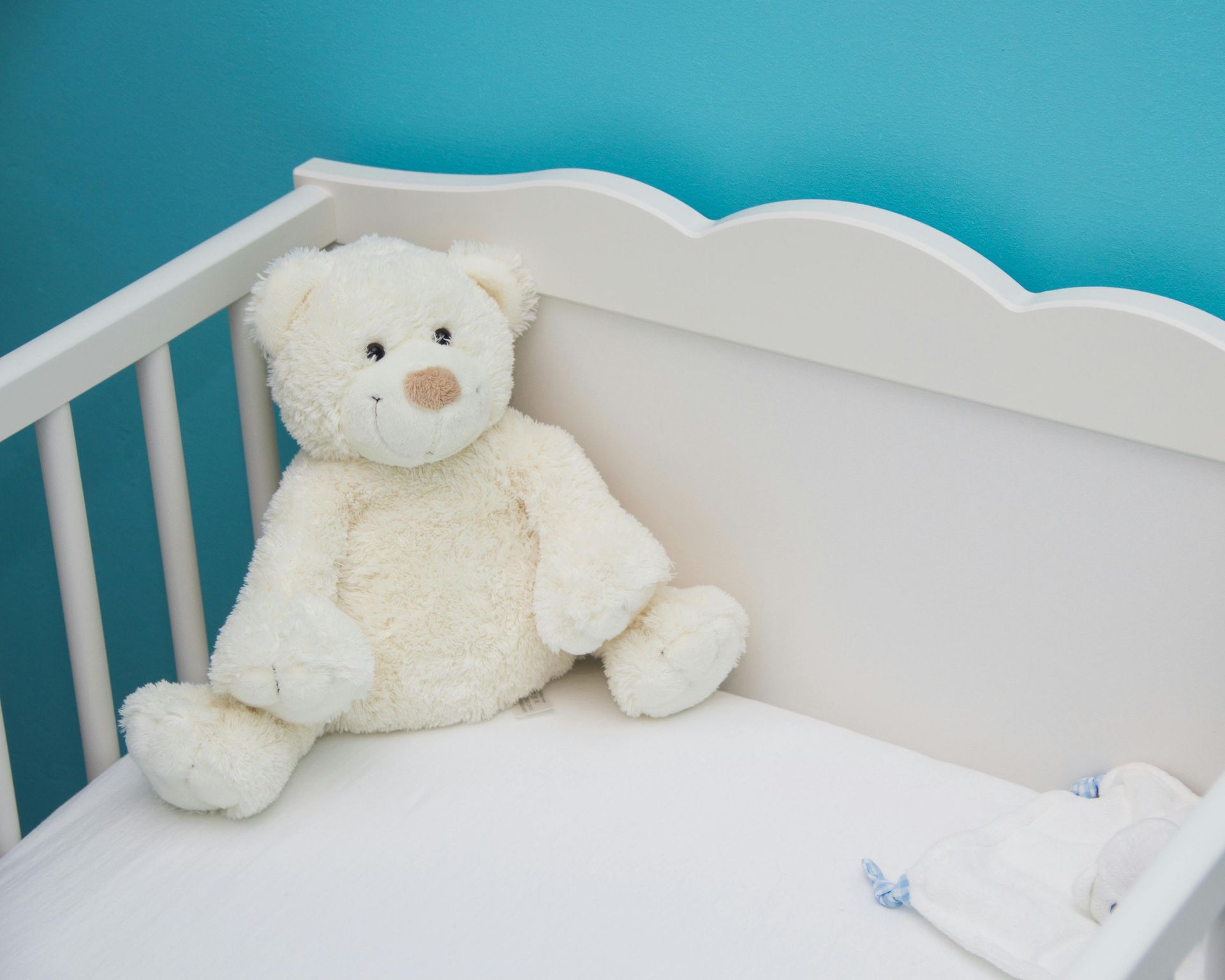 White teddy bear in a white crib, against a blue wall.