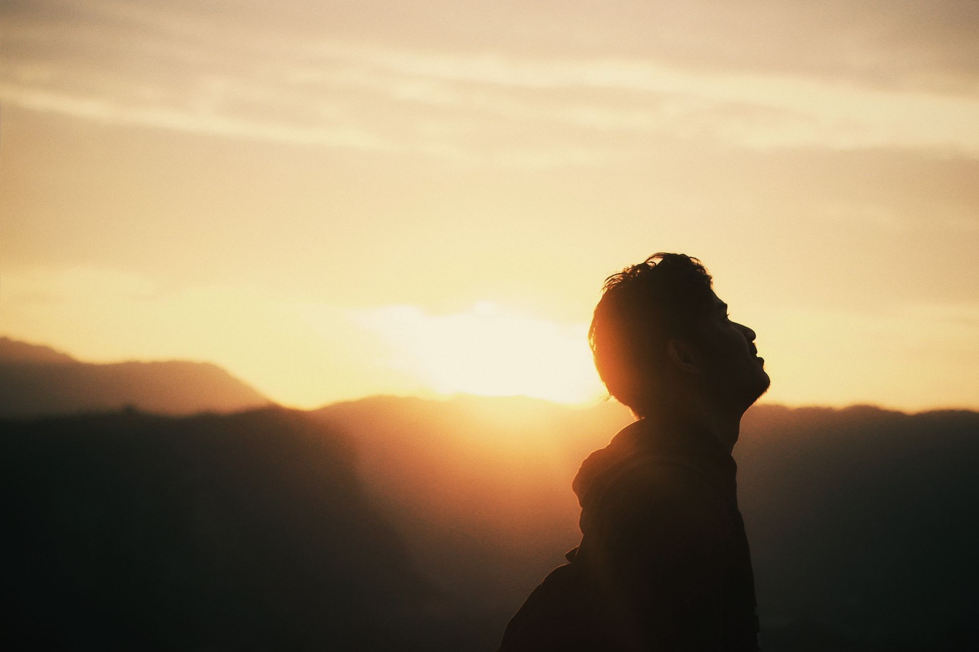 Silhouette of person looking up at a sunset over mountains; warm orange sky.