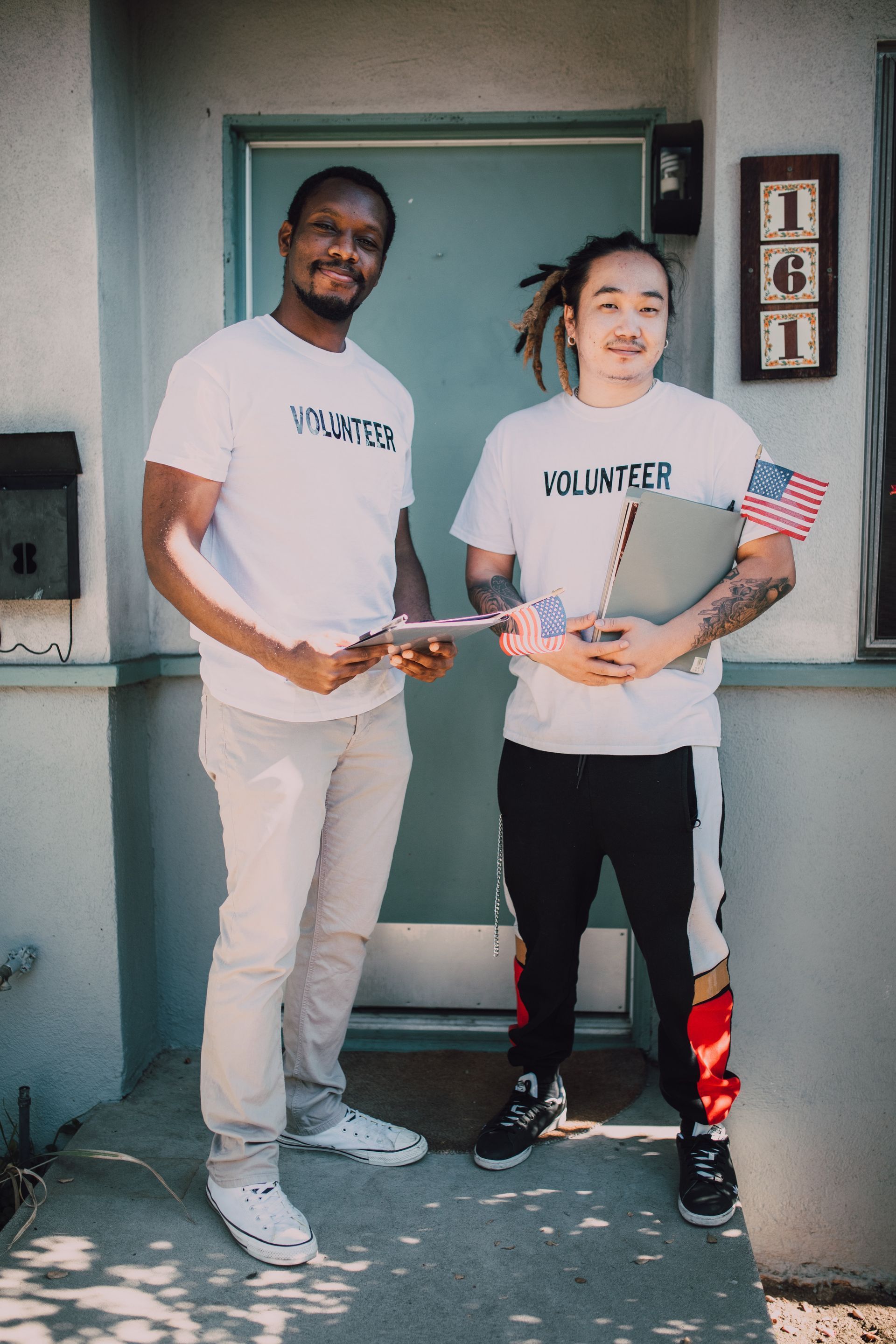 Two volunteers outside a house, holding paperwork and American flags. One man is smiling, wearing a volunteer shirt.