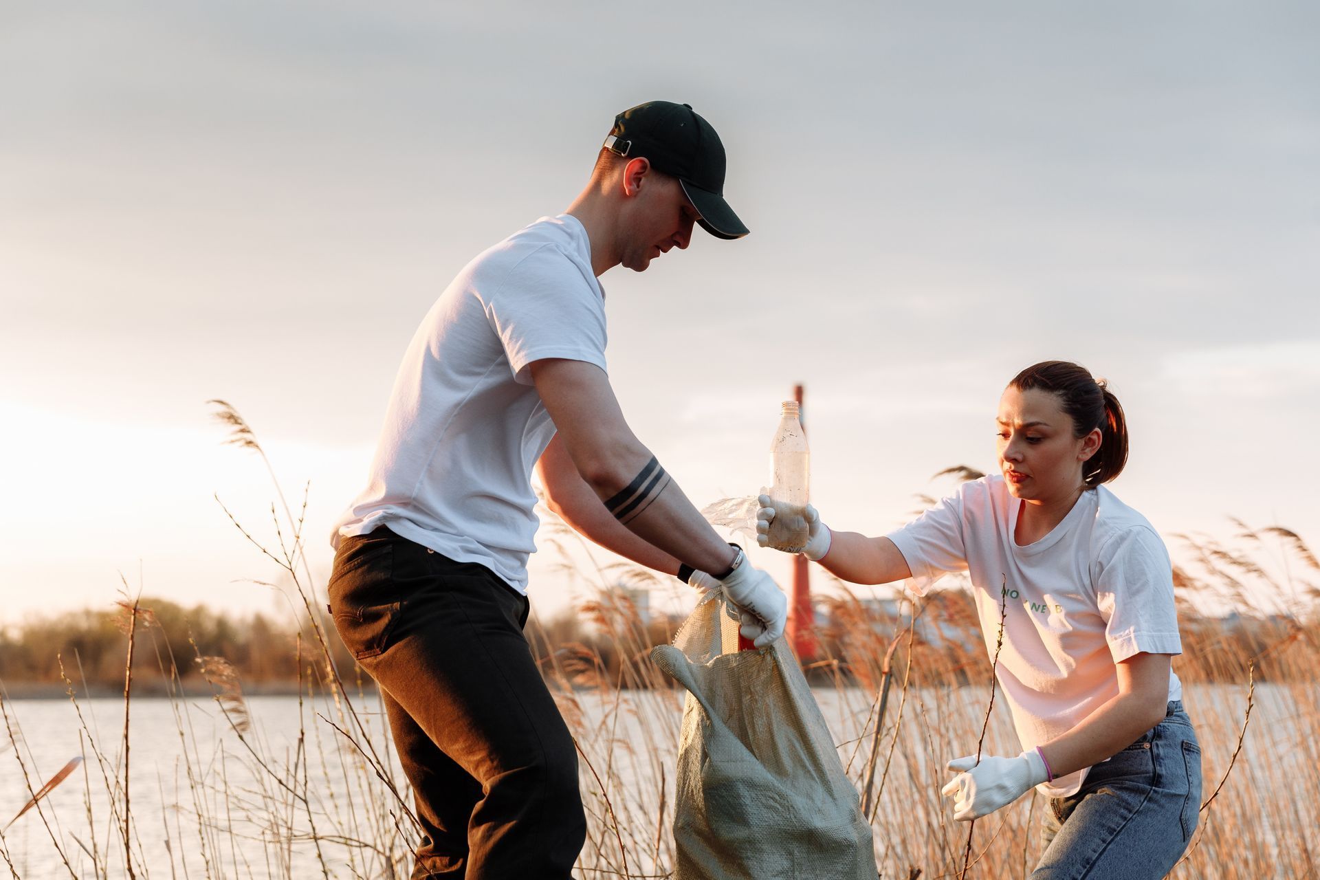 Two people picking up litter by a lake; man in a cap and woman in gloves putting trash into a bag.