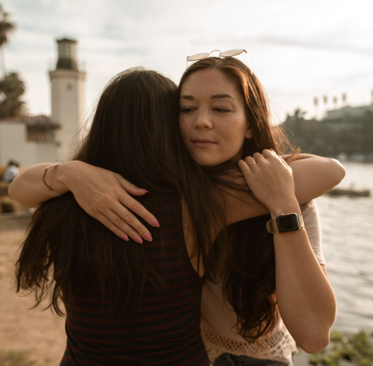Two women embracing by water with a tower and greenery in the background.