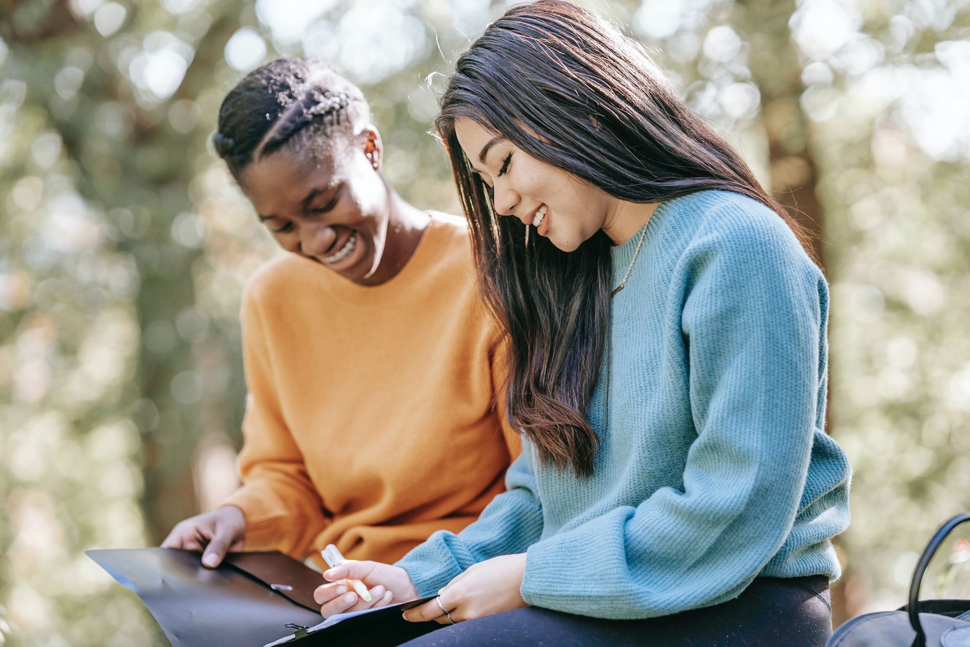 Two young women laughing, writing in a notebook outdoors. One wears orange, the other blue.