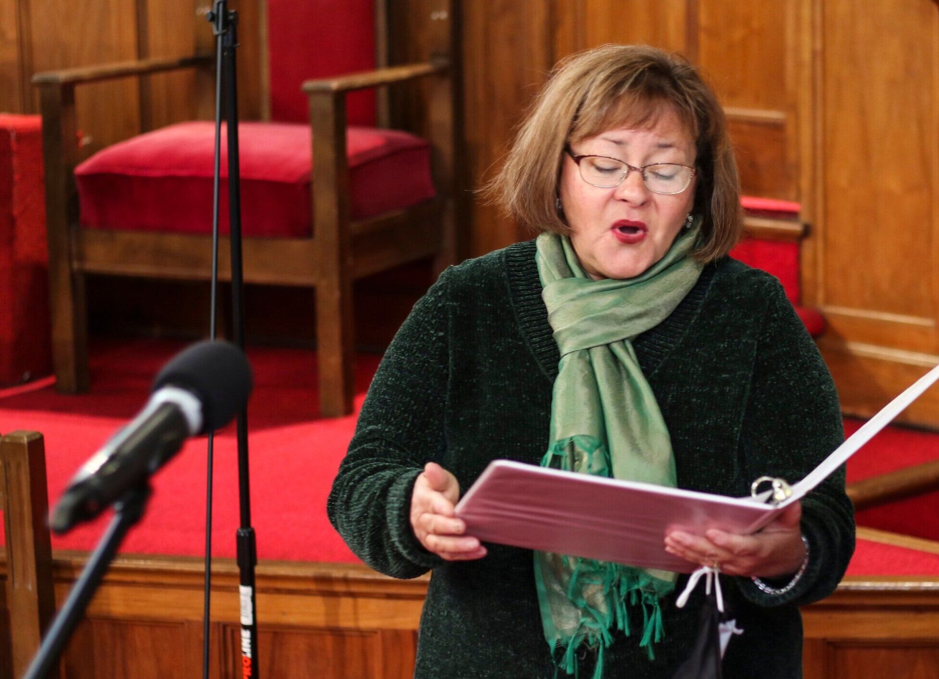 Woman singing into a microphone, holding a book. Wearing green scarf and sweater, standing in a wooden church.