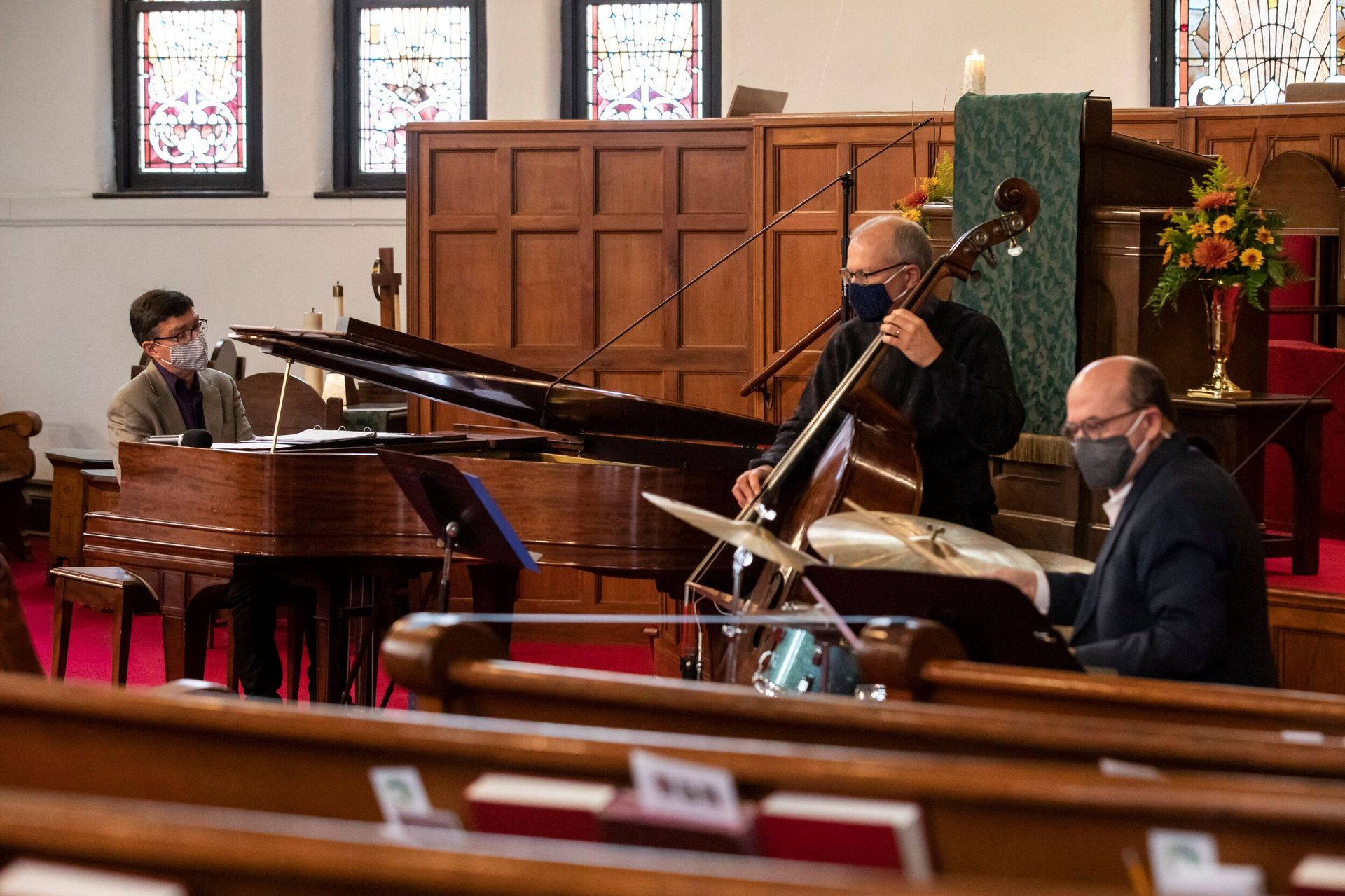 Jazz trio performing in a church sanctuary: piano, bass, drums, all wearing masks.