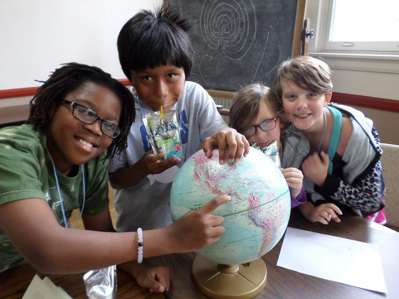 Four children around a globe, pointing at it. Smiling, some wearing glasses, in a classroom.