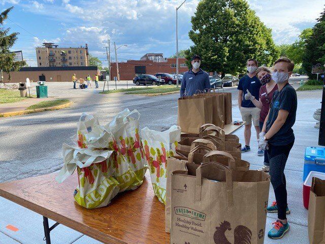 People wearing masks distribute food bags on a table outside. Others work in the background.