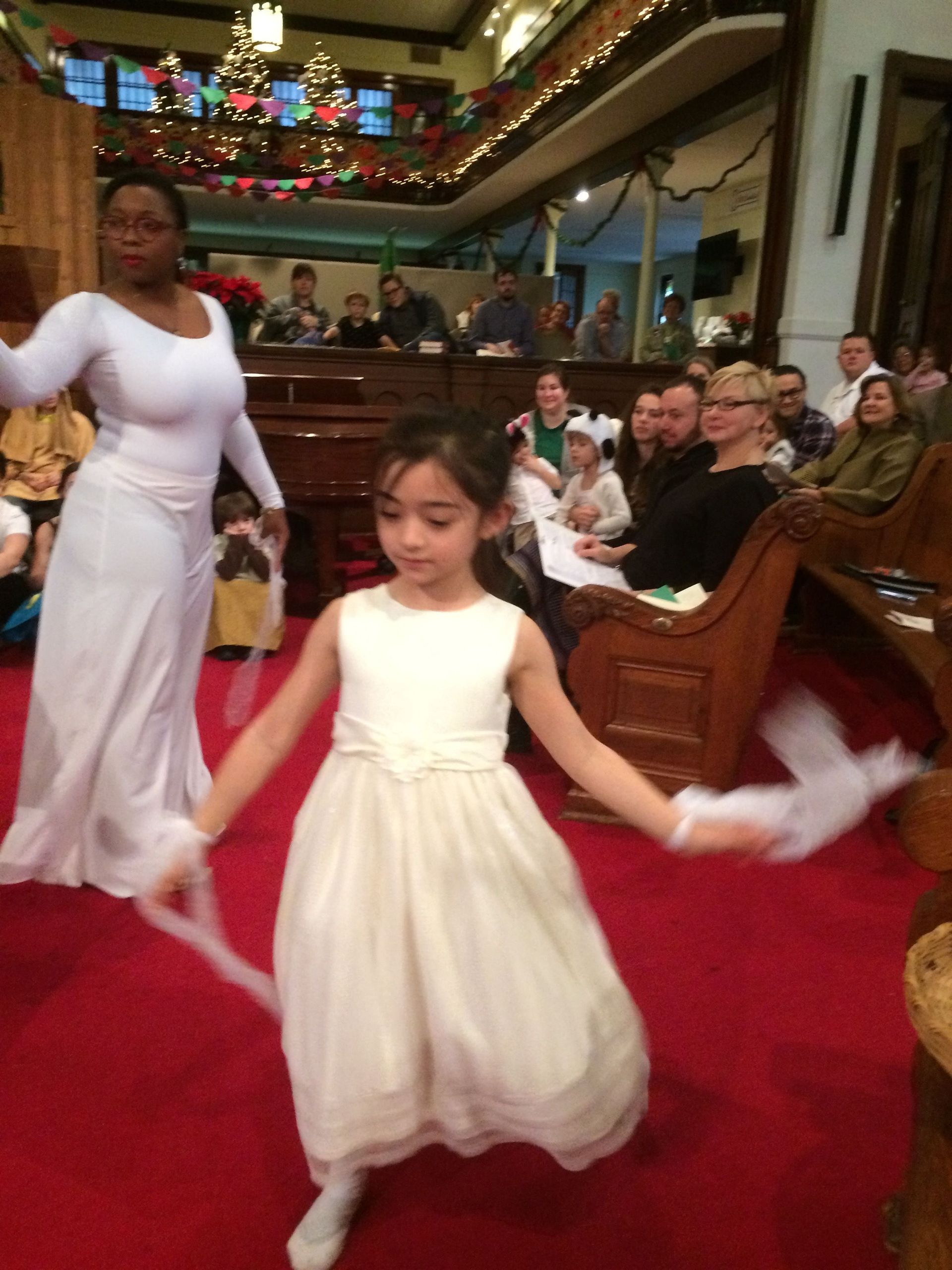 Two women dancing in white dresses in a church. A girl with outstretched arms holds white ribbons.
