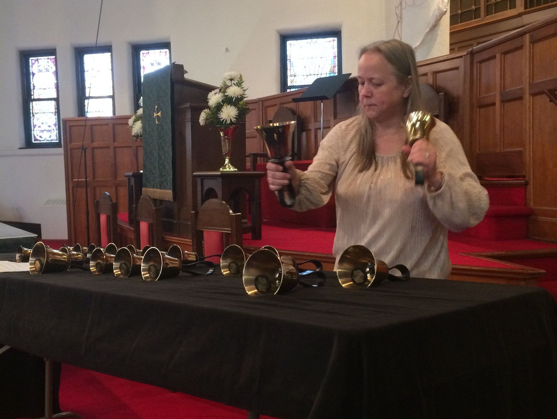 Woman playing handbells in a church; holding two bells and table with multiple golden bells, red carpet.