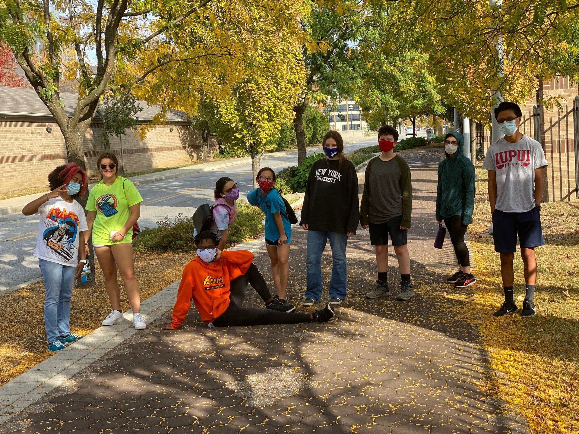 A group of masked students poses outdoors, surrounded by autumn leaves. Some stand, others recline.