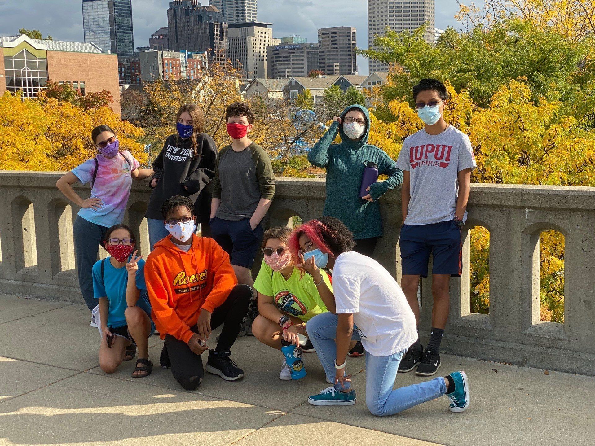 Group of diverse people, masked, posing outdoors against city backdrop, autumn foliage.